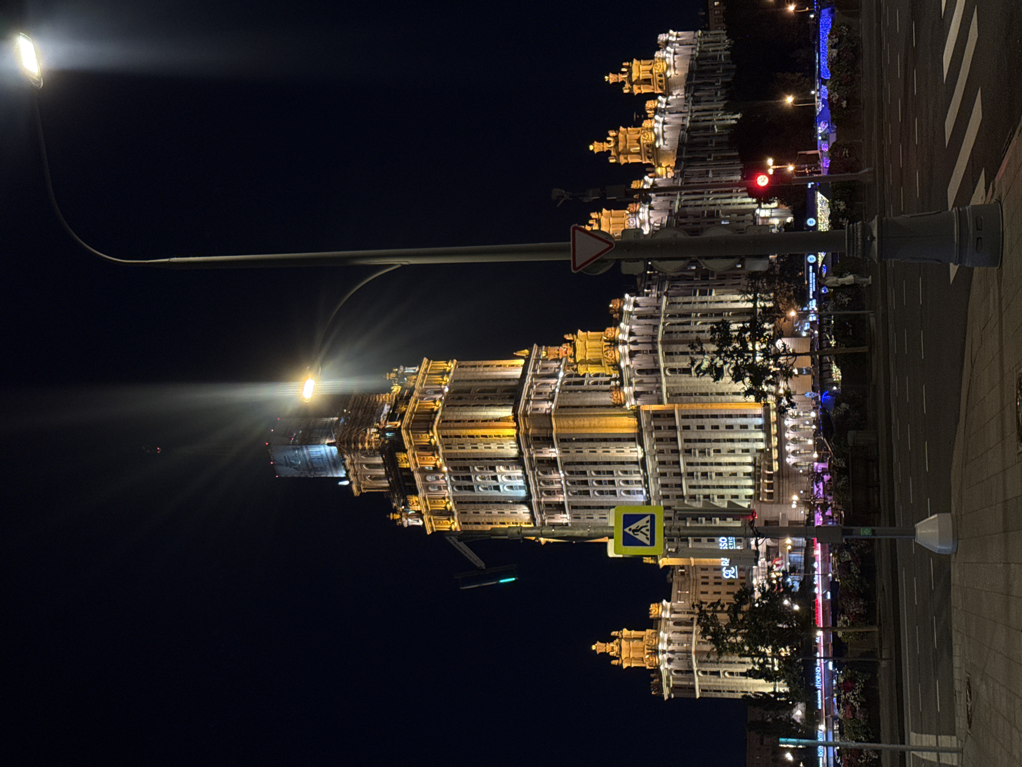 An exterior, eye-level, nighttime shot captures the iconic Stalinist skyscraper, Hotel Ukraina, in Moscow, Russia. The imposing building, illuminated by warm yellow lights, dominates the background. A street light with a bright, sharp beam of light extends from the right side of the frame, casting dramatic rays across the dark sky. In the mid-ground, a pedestrian crossing sign and a traffic light showing green are visible on a pole. The foreground shows a street with white pedestrian crossing lines and a sidewalk. The scene is tranquil, with no visible activity or people, suggesting a late hour.