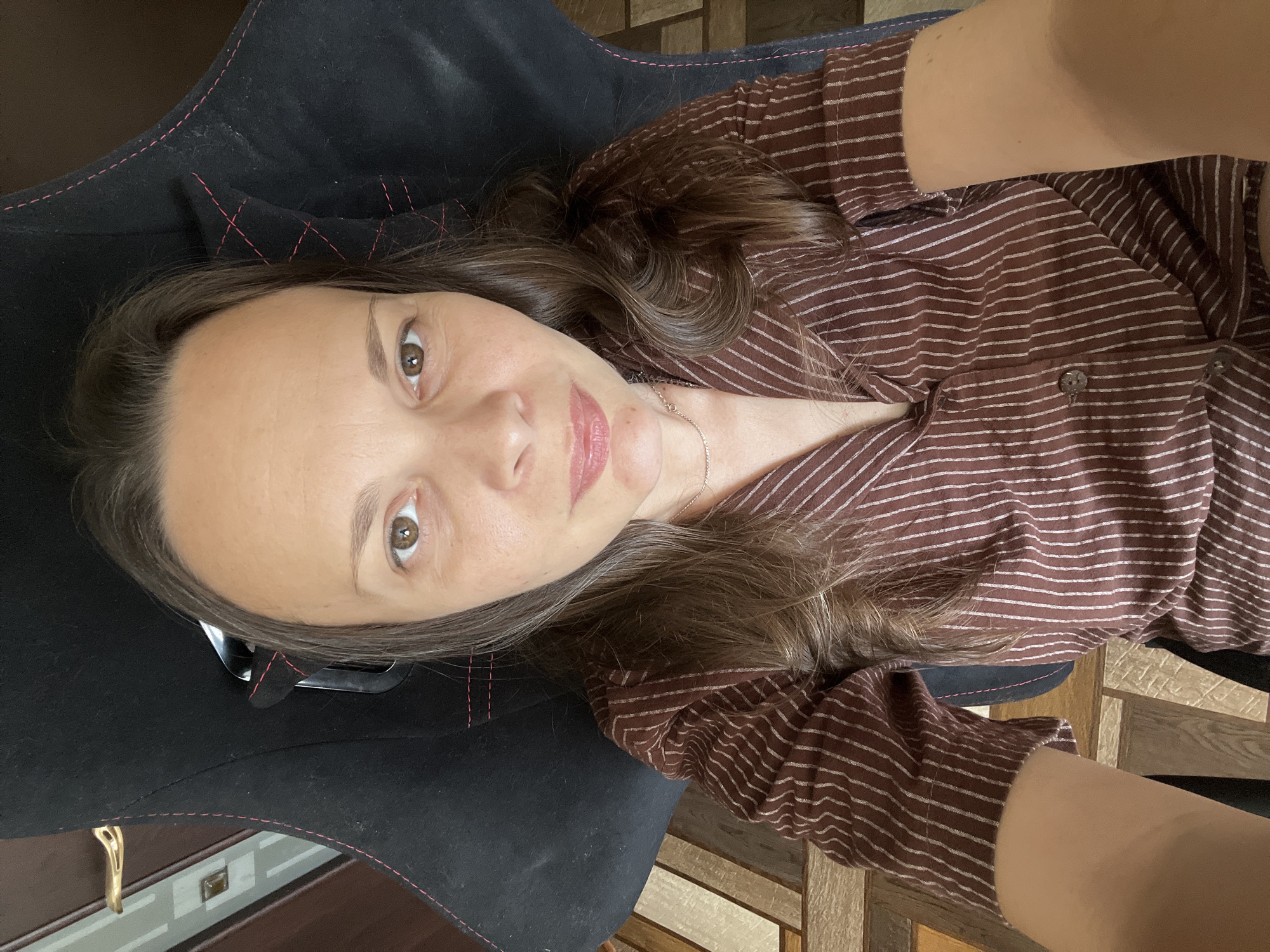 A woman with brown hair is sitting in a black chair. She is wearing a brown and white striped shirt. The background is out of focus but appears to be an indoor setting with wooden flooring and a door. The image appears to be a selfie taken by the woman. There is no indication of the city of Tolyatti, Russia, in the image.