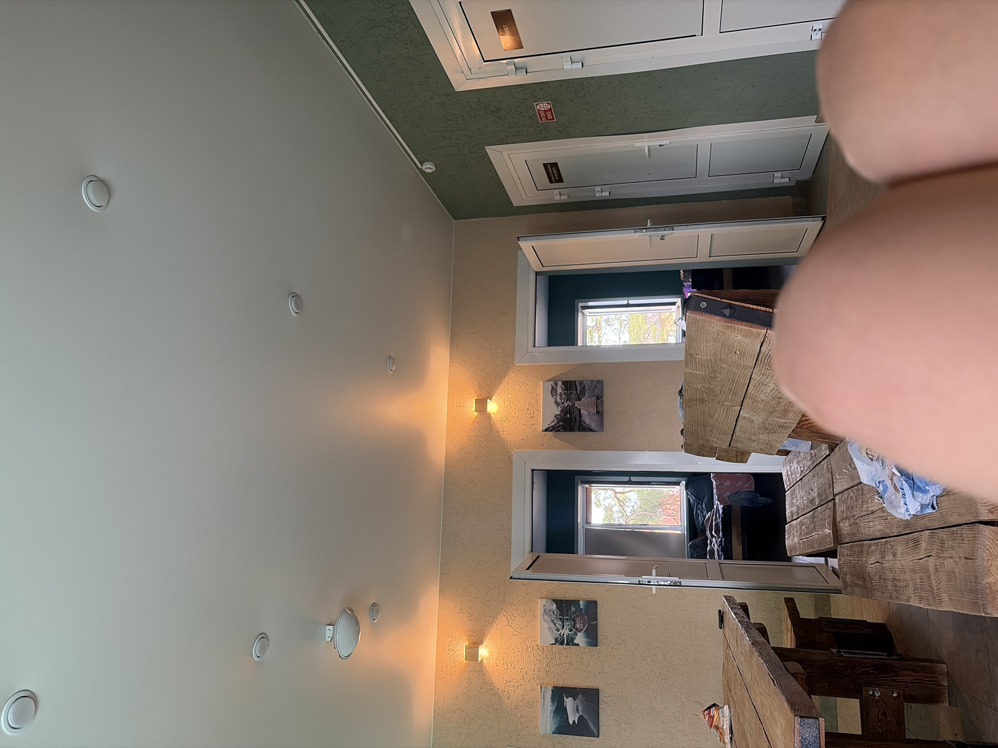 The image shows an interior room with beige walls and a white ceiling dotted with circular light fixtures. The upper ceiling section is green. There are two white doors near the upper part of the image, and two open windows with white frames are visible further down. Between the windows, there are four small, rectangular light fixtures on the wall, along with three pictures of what appears to be nature scenes. A rustic, wooden table and bench set occupies the foreground, though it is partially obscured by a person's hand.