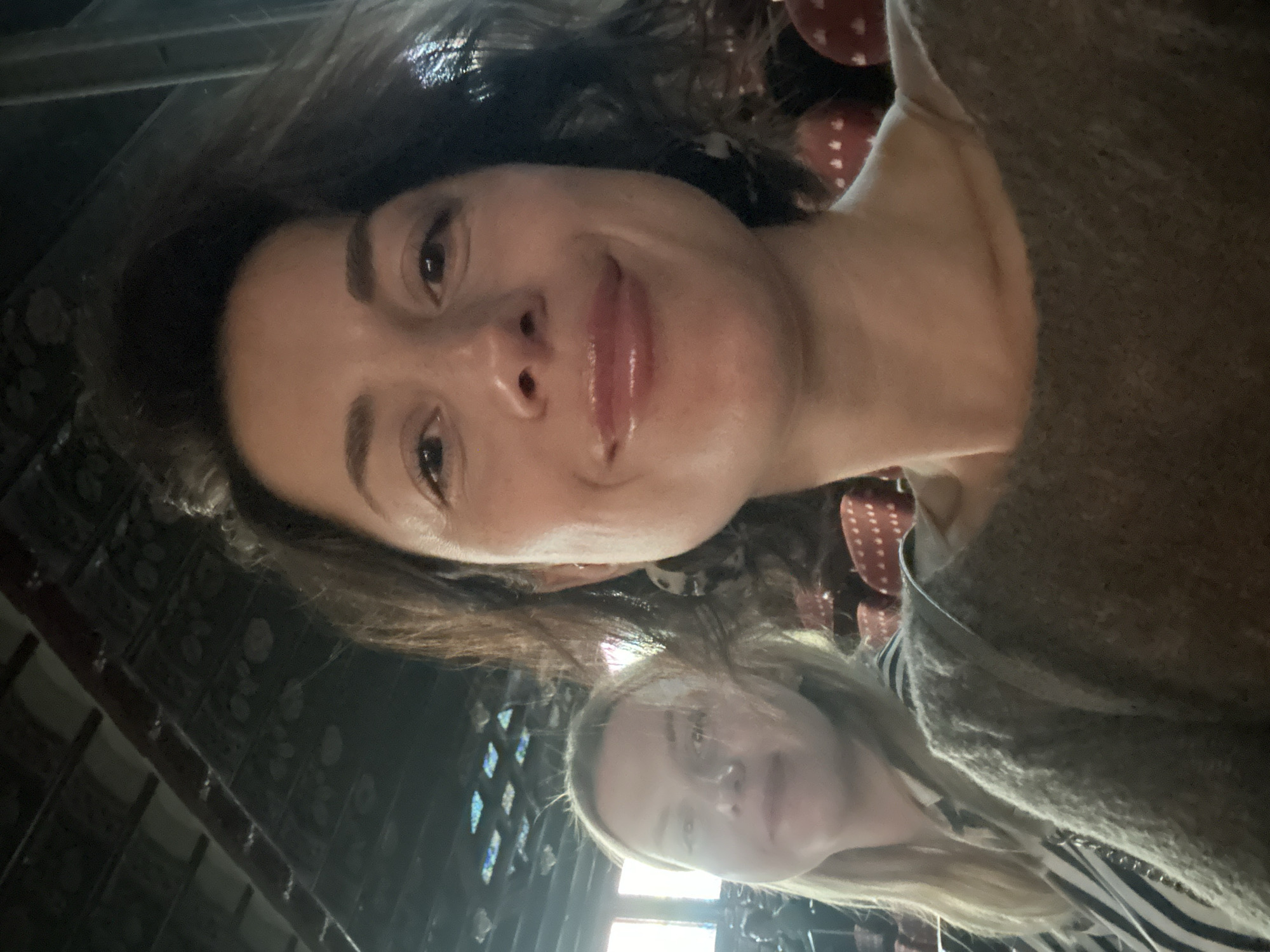 Two women are in the foreground, taking a selfie. The woman closest to the camera has dark hair, fair skin and is wearing a grey sweater. The other woman has blonde hair, fair skin, and is wearing a striped shirt. The location appears to be the Palau de la Musica Catalana in Barcelona, Spain, based on the ornate interior featuring stained glass and decorative elements, suggestive of the modernist architecture of Lluís Domènech i Montaner.