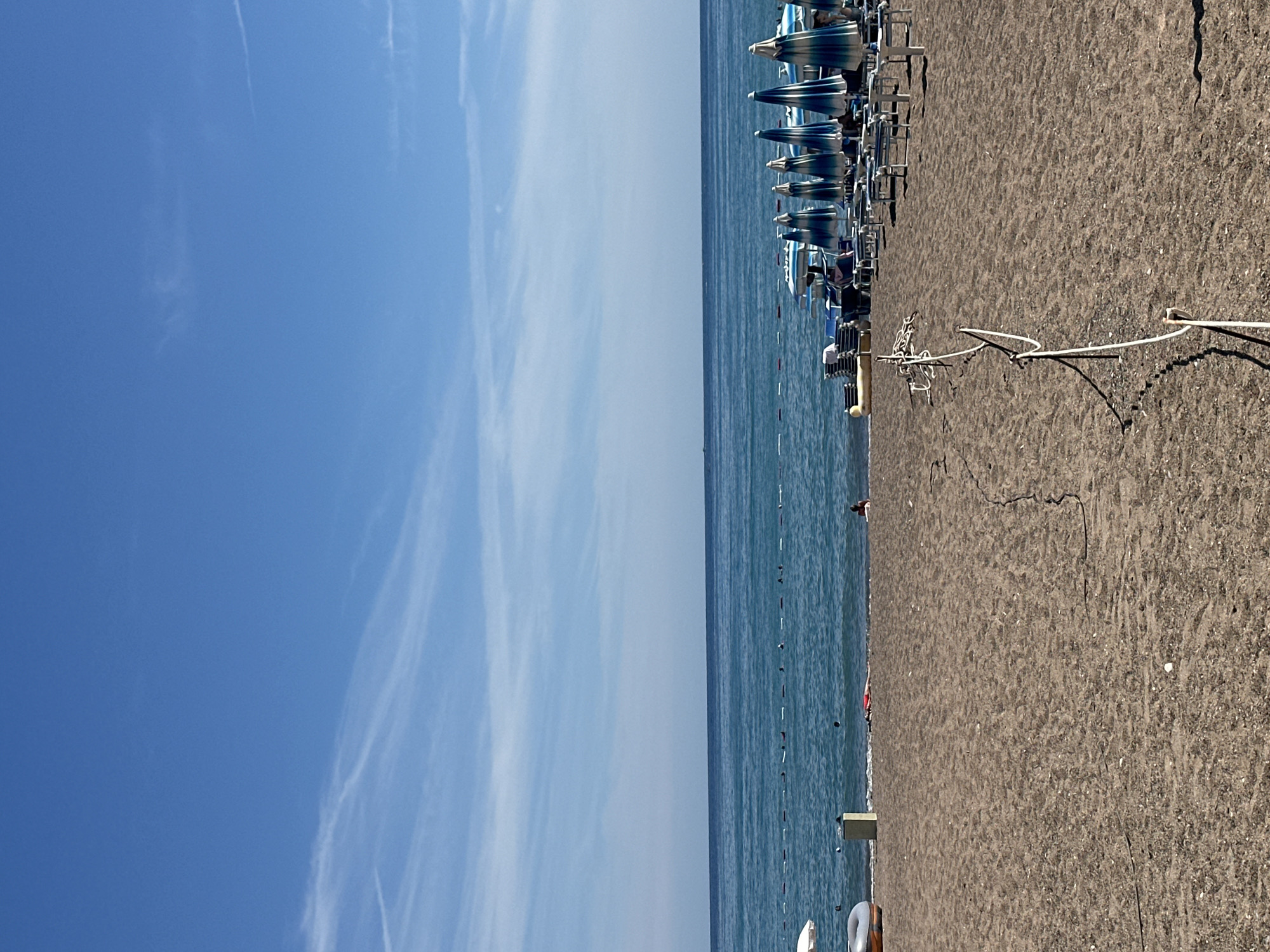 The image depicts a beach scene in the city of Boreti, Montenegro. The sky is a clear blue with wispy white clouds. The sea is a deep blue and calm, with a rope barrier extending into the water. The beach is covered in dark sand, with a row of closed beach umbrellas and lounge chairs set up on the right side of the frame. A few people are visible on the beach and in the water. One person is lying on a towel in the shallow water, and another is sitting on the sand. In the distance, several boats are anchored. The overall impression is one of a peaceful and sunny day at the beach.
