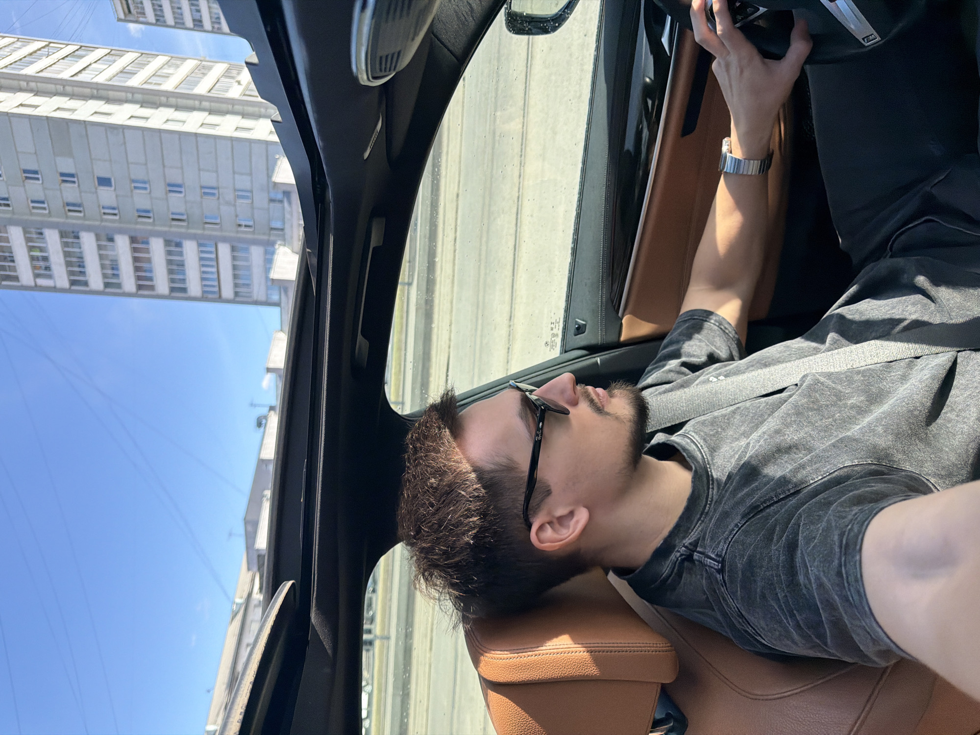 A man with dark hair and a beard, wearing sunglasses and a gray shirt, is seated in the driver's seat of a car with a brown leather interior. He is wearing a wristwatch. The car is on a city street in Krasnoyarsk, Russia, and a tall building is visible through the car's window. The sky is blue.