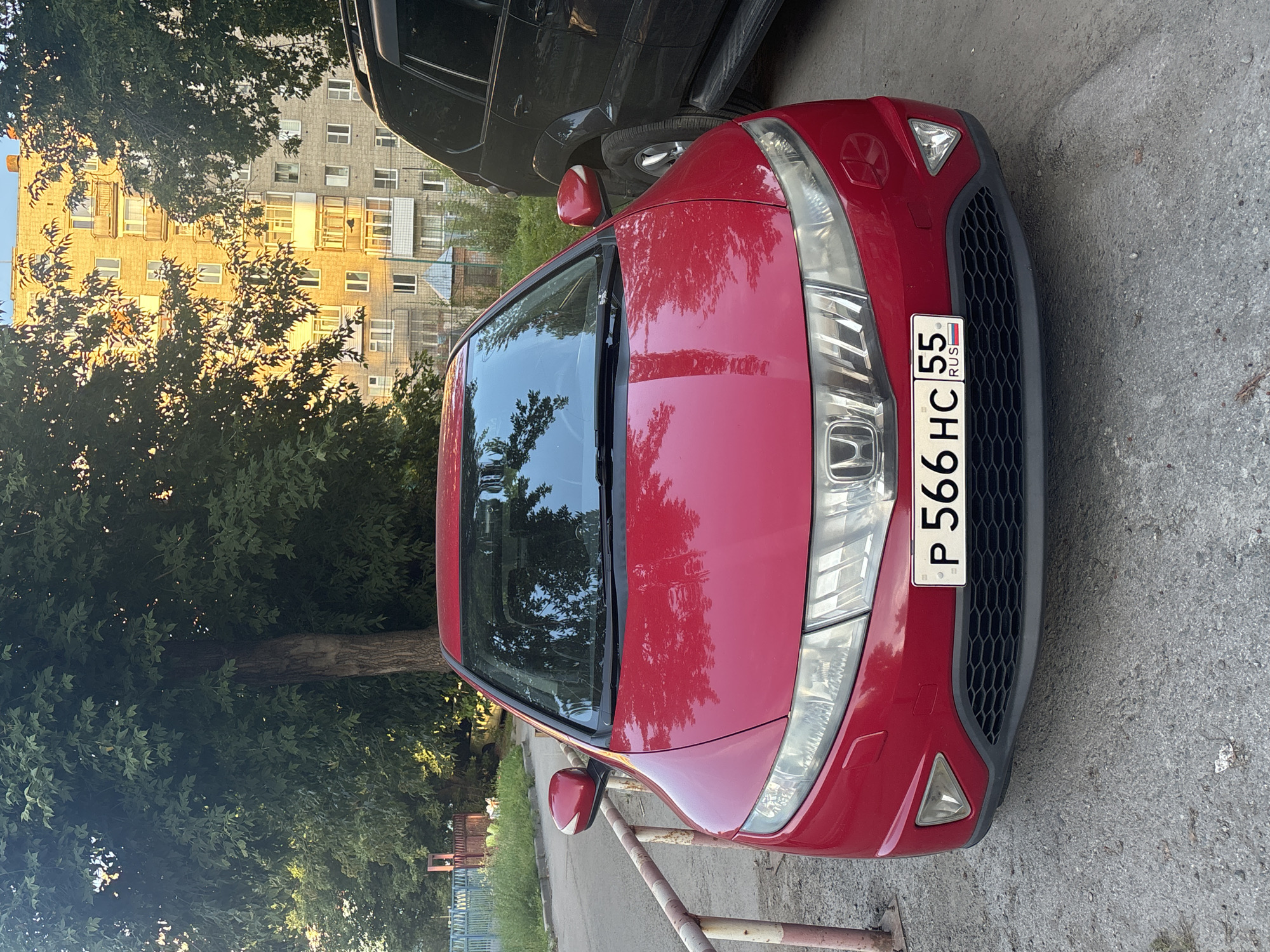 The image shows a red Honda Civic parked on a street in Novosibirsk, Russia. The car is facing the camera, and its license plate reads "P 566 HC 55 RUS". Behind the car, there is a tall apartment building and several trees. The sun is shining, casting a warm glow on the building. The street appears to be paved with asphalt and is lined with a metal railing. A black SUV is parked to the right of the Honda Civic. The environment is urban, with elements of residential architecture and natural vegetation.