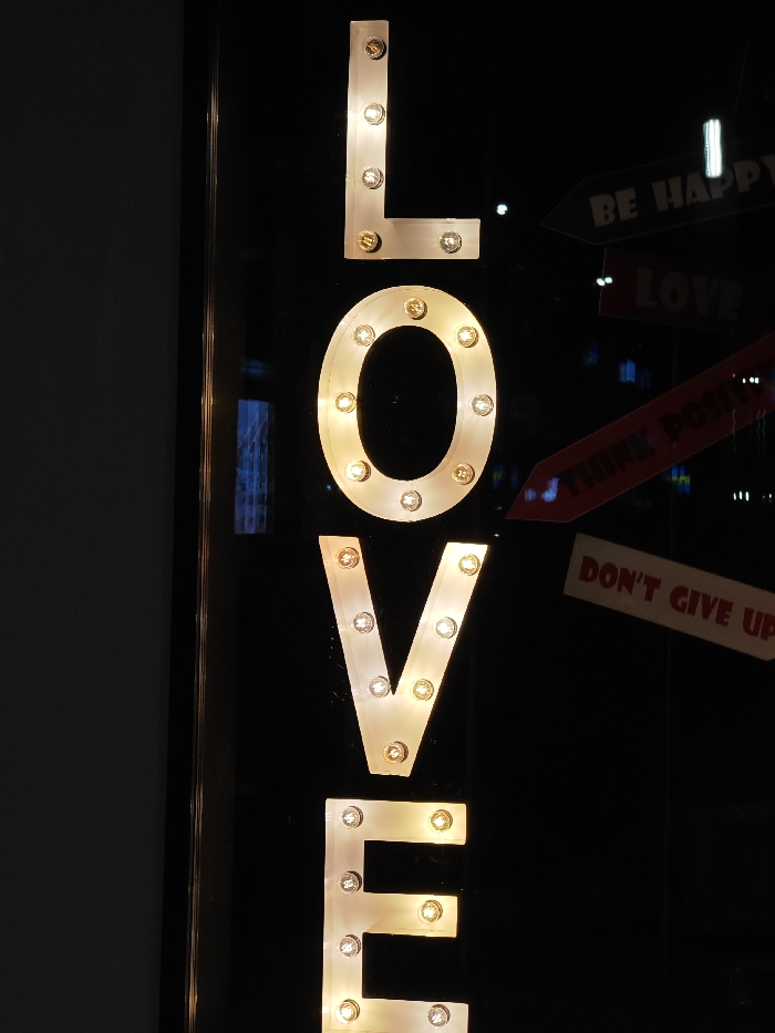 A vertical marquee-style sign spelling "LOVE" is centrally displayed against a dark, reflective background. The individual letters, light-colored and outlined, each contain multiple embedded warm-toned incandescent bulbs that illuminate them. Reflections are prominent on the right side of the frame, revealing portions of three arrow-shaped signs with text: "BE HAPPY" is visible in the upper right, a partially obscured red-brown sign likely reading "THINK POSITIVE" is below it, and "DON'T GIVE UP" is discernible in the lower right. No individuals, direct actions, or interactions are visible within the frame. This scene is located in the city of Shumen, Bulgaria.