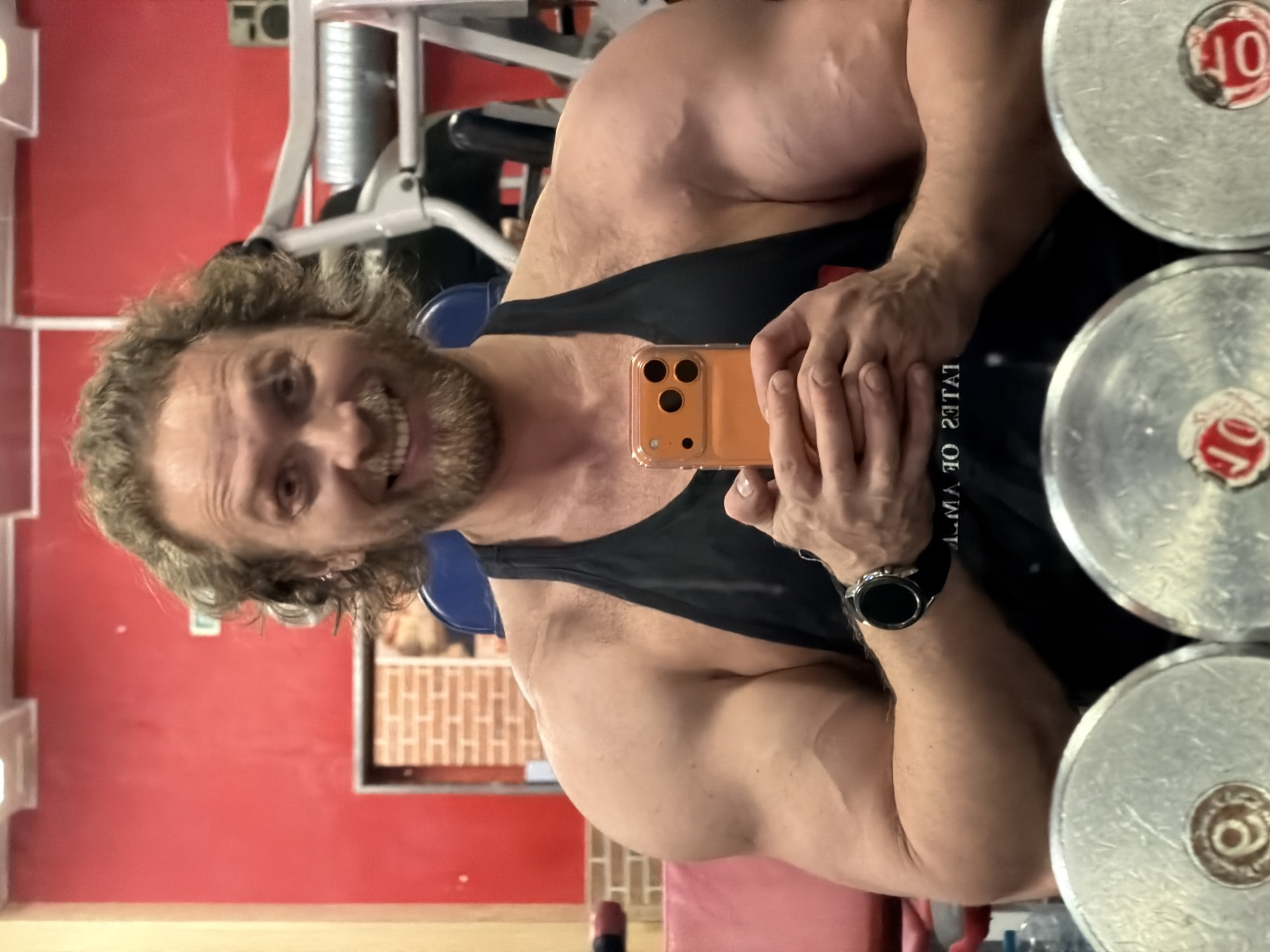 A male individual with curly blonde hair and a beard is depicted from the chest up, positioned directly facing the viewer. He wears a black tank top, revealing prominent musculature in his shoulders and arms, with visible veins on his forearms. A dark smartwatch is on his left wrist. He is smiling broadly, exposing his teeth. Both hands clasp an orange-cased smartphone, which he uses to capture a mirror selfie. The reflection of his shirt on the phone's screen partially shows "ОФ САНКТ-ПЕТЕРБУРГ" (OF SAINT PETERSBURG), indicating the location is in Saint Petersburg, Russia.
The background, visible around and behind the man, is a gym interior. Features include a bright red painted wall and sections of exposed reddish-brown brickwork. Various pieces of exercise equipment are present: a blue-padded machine is behind his left shoulder, and a white-framed machine with silver components is behind his right shoulder. In the immediate foreground, partially visible below his arms, are the chrome tops of two large dumbbells or weight plates. Overhead, white rectangular lighting fixtures are visible on the ceiling. The environment is well-lit.