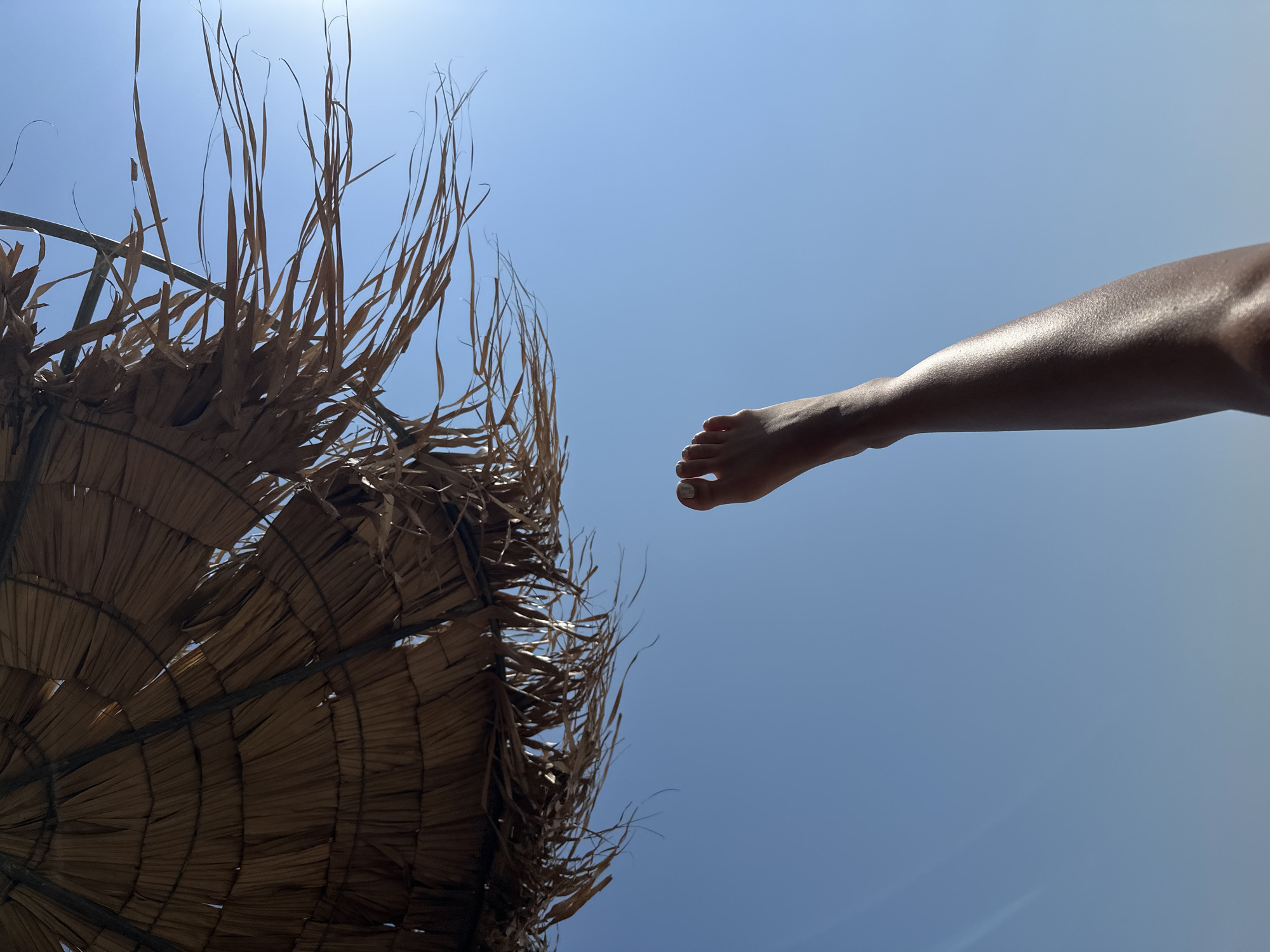 This is a low-angle, full shot of a person's leg and foot extending upwards against a clear blue sky. A portion of a thatched umbrella is visible in the upper left corner of the frame, casting shadows. The person's skin appears tanned and is catching the sunlight, creating highlights. The scene is set on Ofrynio Beach in Greece.