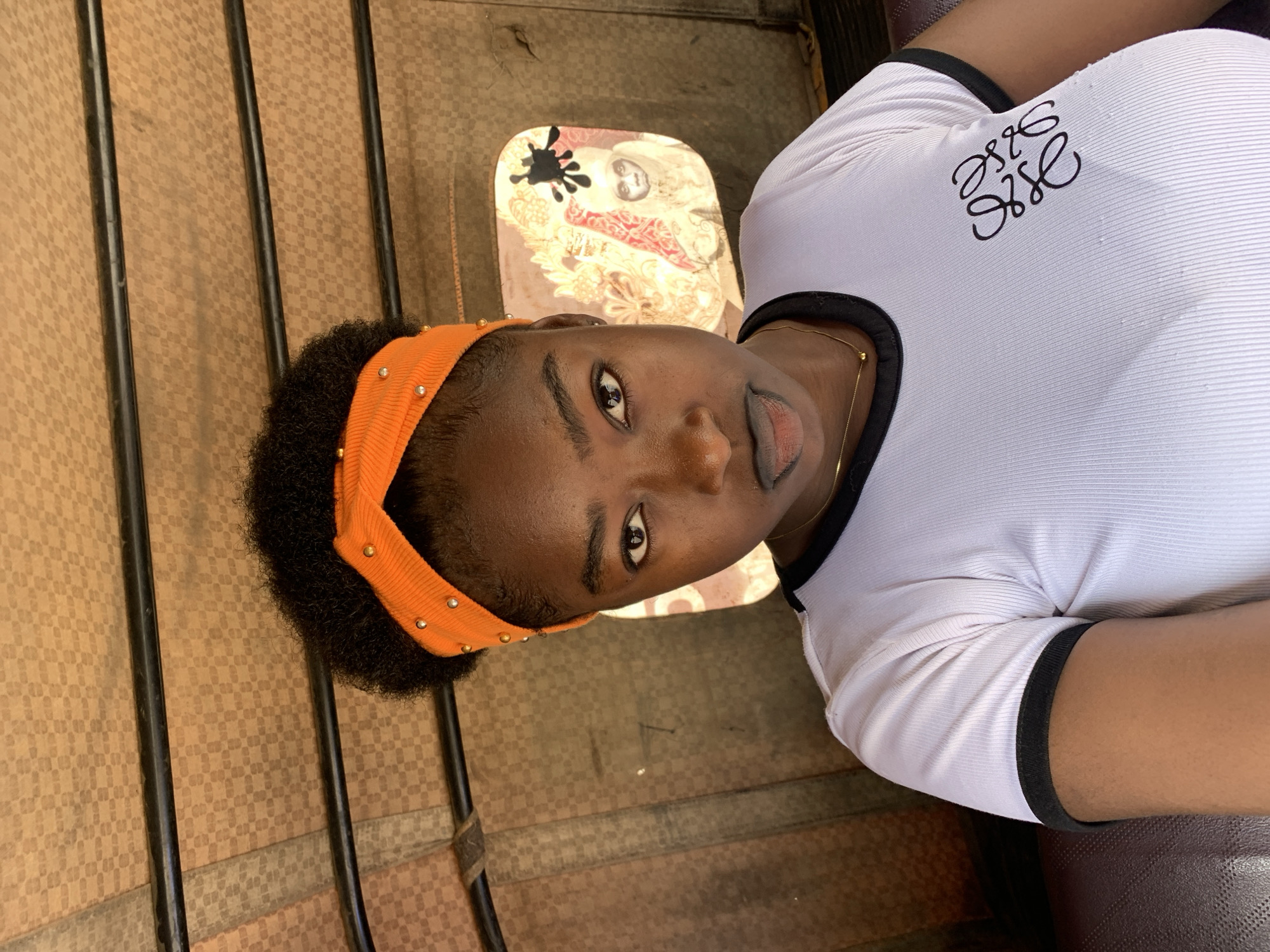 A young woman is pictured from a high-angle view inside a vehicle in New Stadium, Nigeria. She wears an orange headband with small embellishments and a white t-shirt with a black logo and trim. The interior of the vehicle shows a decorative upholstery panel above her and barred window frames. The overall lighting is natural daylight.