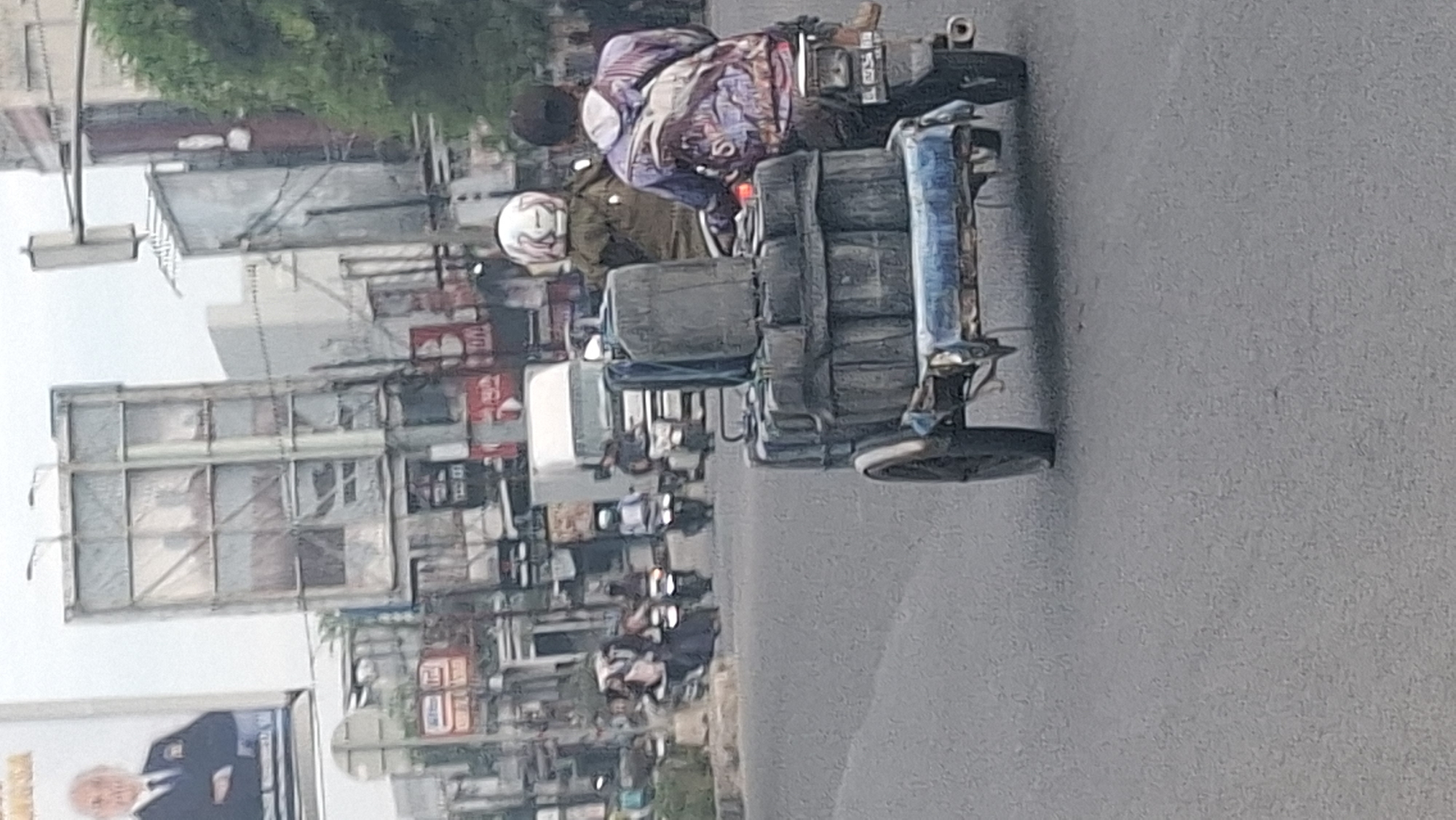 This is a medium-angle, eye-level, outdoor photograph of a street scene in Lhokseumawe, Indonesia. The image focuses on a motorcycle with a trailer carrying several large, black, plastic containers. Two people are riding the motorcycle; the driver is wearing a white helmet with pink and white markings, and the passenger is wearing a black helmet and a colorful, patterned jacket. The motorcycle and trailer are in the foreground and center of the image, moving towards the right.
In the middle ground, there is a white truck, several other motorcycles, and some pedestrians. The pedestrians on motorcycles appear to be mostly male, and some are wearing helmets. The environment is urban, with buildings and traffic signals visible in the background. There are also several billboards, one of which shows a portrait of a man in a suit and tie. The sky is overcast, and the lighting suggests it is daytime. The overall scene depicts a busy street with traffic and commerce.
