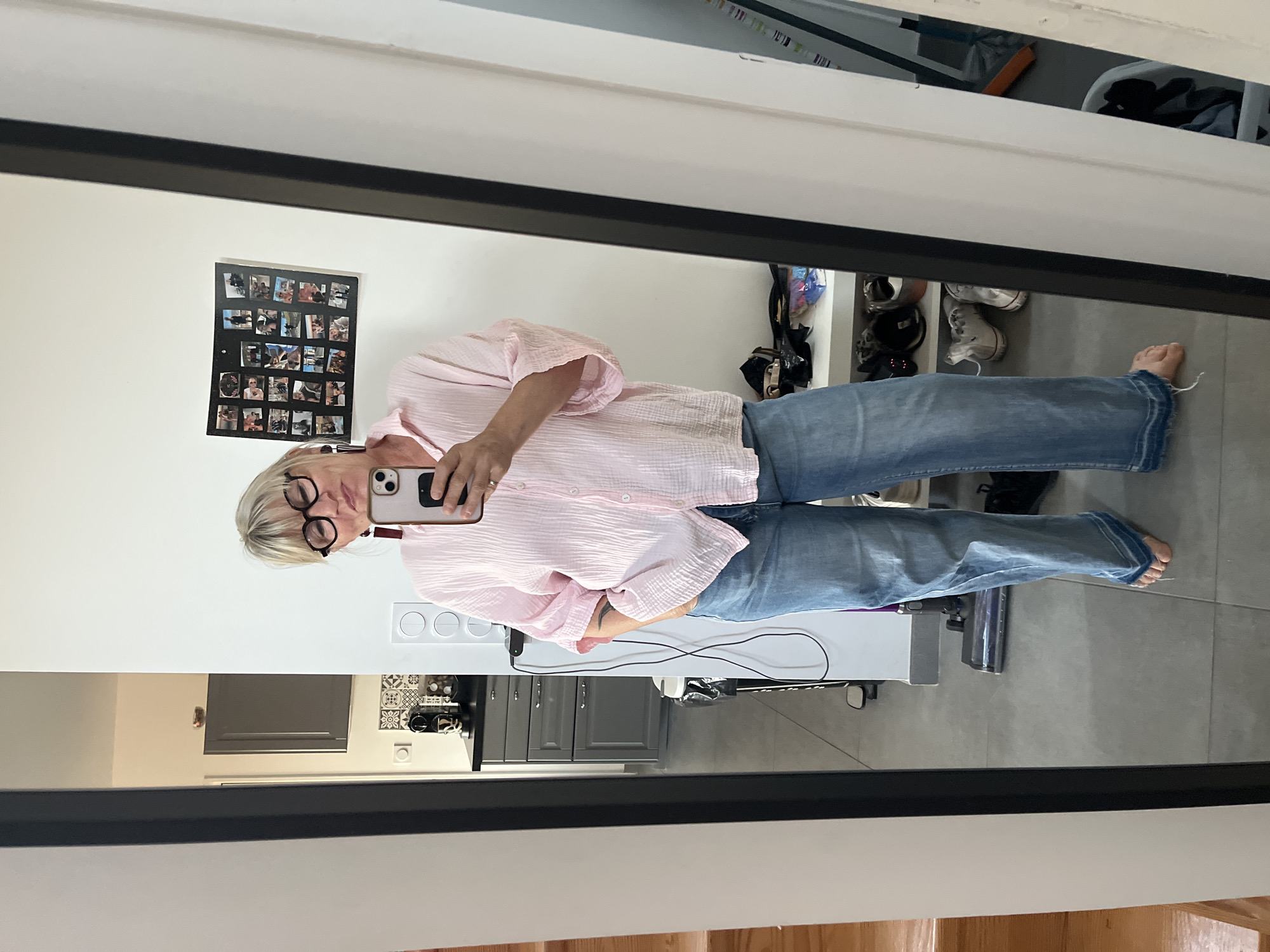 The image depicts a woman in a mirror selfie. She has short blonde hair, wears black-rimmed glasses, a light pink button-down shirt, and blue jeans. She is holding a smartphone in her right hand to take the picture. The setting appears to be inside a house, with a kitchen visible in the reflection, featuring gray cabinets and a tiled backsplash. There is also a vacuum cleaner and a white storage unit with shoes. On the wall behind the woman in the reflection, there's a collage of photos. There is no clear information about the location being in the city of Chambœuf, France.