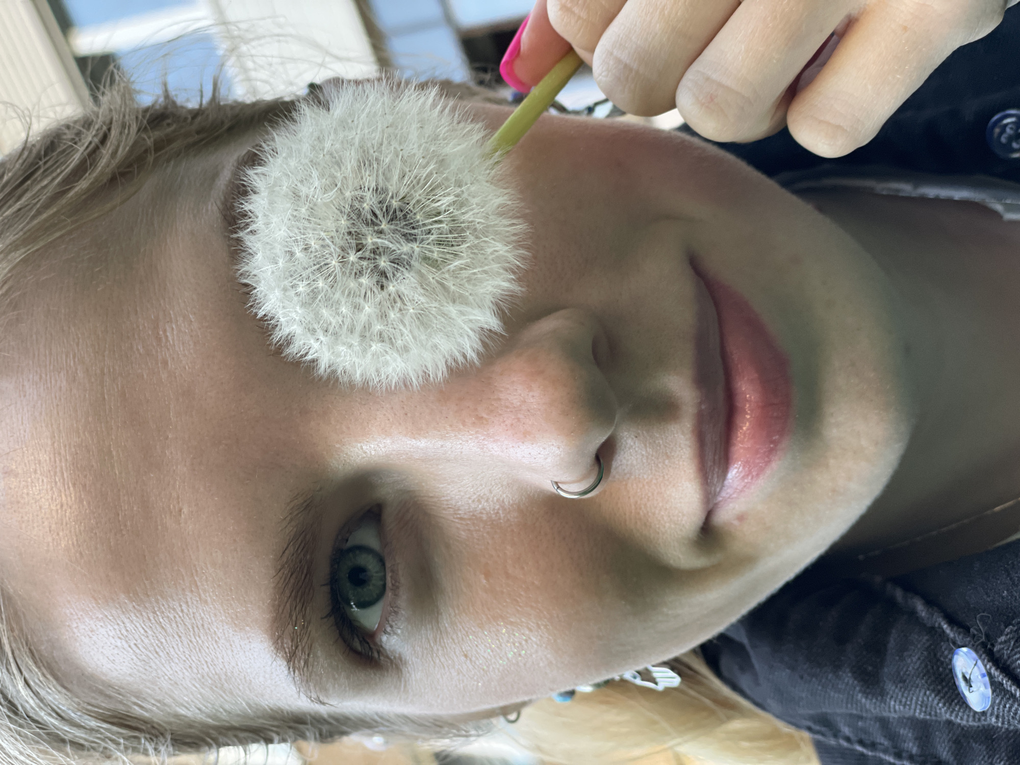 A close-up shot of a woman with fair skin and light-colored hair, holding a dandelion seed head. She has a nose ring in her left nostril, and pink nail polish on her finger. Her eye color is green, and her lips are pink. She is wearing a dark-colored denim jacket. The background is out of focus. It is not possible to verify the location, Izhevsk, Russia, based on the image.
