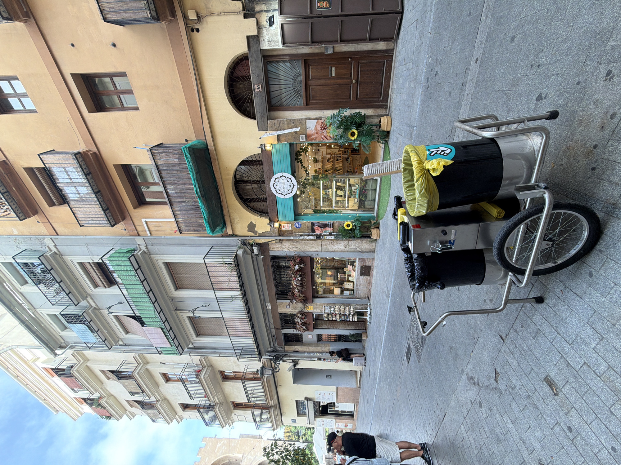 A street scene in an urban environment features a narrow cobblestone street lined with multi-story buildings. In the foreground, a silver and black cleaning cart with two large trash bins, one lined with a yellow bag and adorned with a peace sign sticker, is positioned on the street. A person wearing a black t-shirt and white shorts stands to the left of the cart, facing away from the camera. Further down the street, two shops are visible. The shop on the left has a display of items outside, while the shop on the right, with a teal awning and the name "Mandala" in a circular logo, showcases products within its well-lit interior. A dark wooden door is situated to the right of the "Mandala" shop. The buildings exhibit balconies with wrought-iron railings and windows, some covered with bamboo blinds. The sky above is partly cloudy.