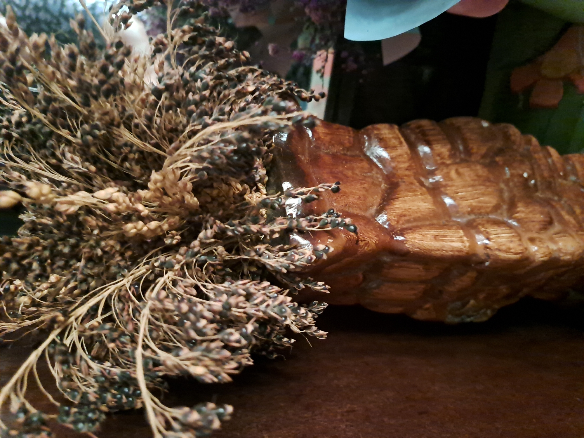 A close-up shot shows a bouquet of dried seed stalks in a textured, brown ceramic vase. The vase has vertical ridges that give it a carved appearance, and a glossy finish. The seed stalks are a mix of brown and dark tones, with many small, round seeds clustered together. The background is a dark, textured surface, likely a wall, which is a warm brown color. To the right of the vase, a blurry light blue object is partially visible, possibly another vase or decorative item. The lighting is dim, creating shadows and highlighting the textures of the vase and dried plants. The scene appears to be indoors, possibly in a home or a shop.