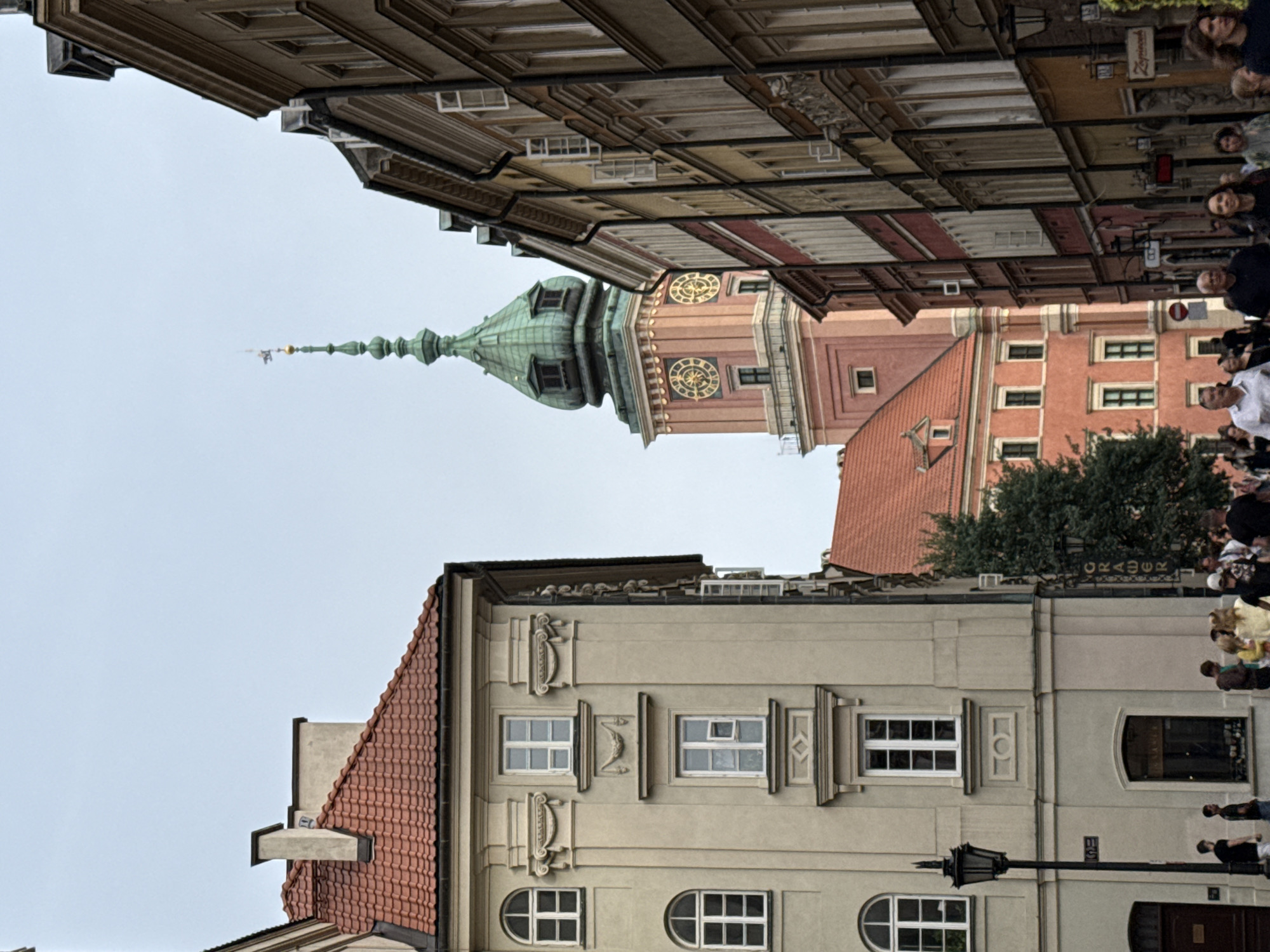 The image depicts a street scene in Warsaw, Poland, with buildings and people. The perspective is unusual, with the buildings appearing to extend upwards from the bottom of the frame and the sky in the upper portion, suggesting the photo might have been taken looking up or is intentionally rotated.
Dominating the middle ground is a distinct architectural feature, likely a clock tower, characterized by its green, pointed spire and a reddish-brown body with large, circular clocks featuring Roman numerals. Below this, a long, multi-story building with a red-tiled roof is visible, characterized by numerous windows and decorative architectural elements. To the left, another building with a similar red-tiled roof is partially visible.
In the foreground, a lighter-colored building displays several windows of various shapes and sizes, including rectangular and arched ones. Decorative mouldings and pilasters adorn its facade. A black lamppost stands prominently in front of this building.
People are scattered throughout the scene, particularly in the upper right portion of the frame, suggesting a public space. They are dressed in casual attire, and their presence indicates activity or a gathering. One group appears to be observing something, while others are walking.
The sky is overcast, suggesting a cloudy or possibly rainy day. The overall impression is of an urban environment with historical architecture.
Visible text includes "CRACOWIA" on a sign above a doorway, indicating a potential connection to Kraków or a business named after it.