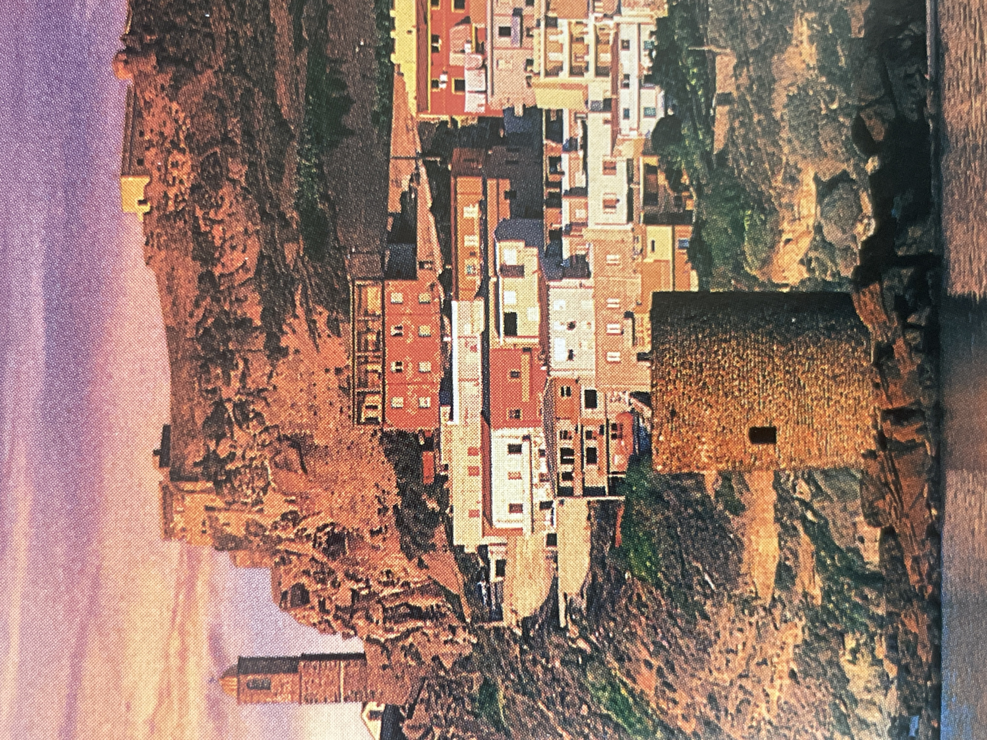 This image depicts a coastal town with multi-story buildings clustered on a rocky hillside. A prominent stone tower stands in the foreground, partially surrounded by water. In the background, a larger stone structure or fortification sits atop the hill, with a tall, slender tower to its left. The sky exhibits a gradient of purple and orange hues, suggesting a sunset or sunrise. The buildings are a mix of colors including red, white, and yellow, with visible windows and balconies. There are no people or vehicles clearly visible in the scene. The overall impression is one of a historic, picturesque European coastal settlement. The location is identified as being in the city of Sofia, Bulgaria.