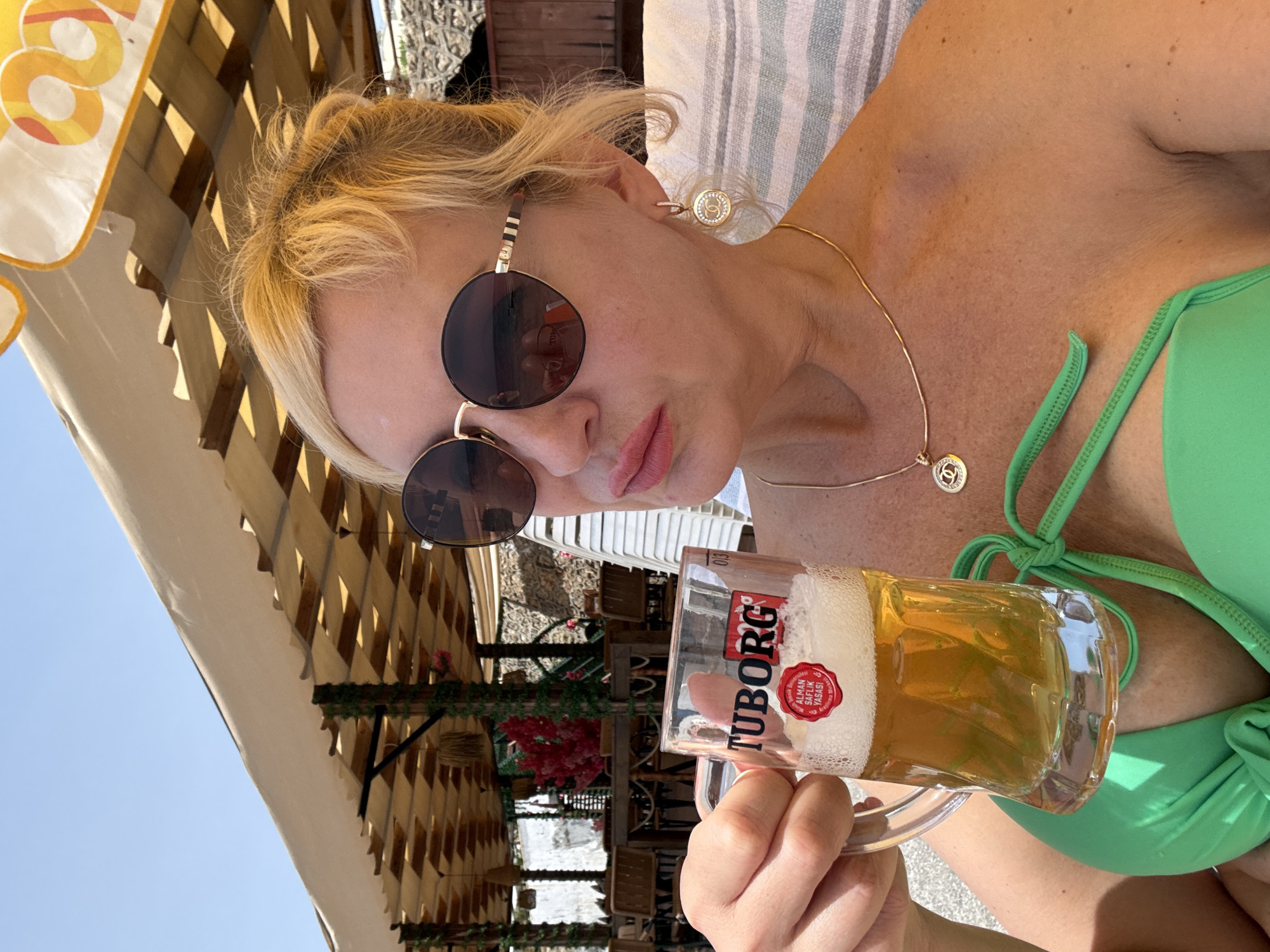 A woman with blonde hair is holding a mug of Tuborg beer. She is wearing round sunglasses, a gold necklace, and a green bikini top. The background appears to be an outdoor cafe or beach setting with tables, chairs, and awning-like structures. The exact location and city are not identifiable from the image.