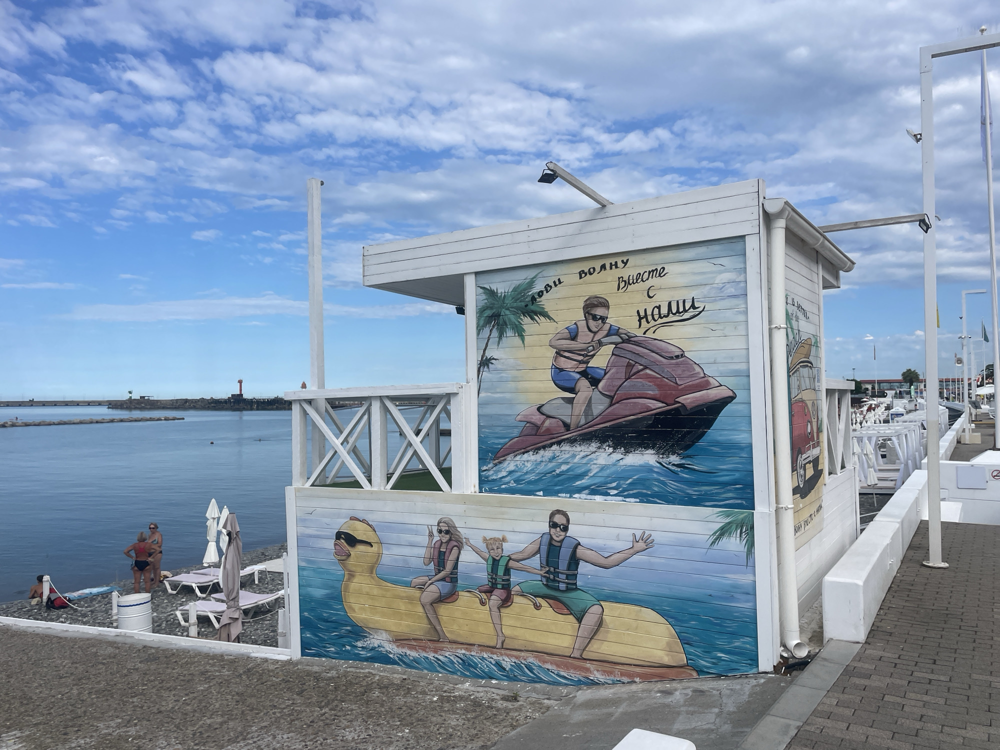 The image depicts a beach scene in Sochi, Russia, with a white wooden structure adorned with colorful murals. The murals feature people enjoying water activities. One mural shows a man on a jet ski, with the Russian text "Лови волну Вместе с нами" (Catch the wave with us). Another mural depicts a family of three, including a child, riding on a yellow inflatable duck, all wearing life vests and sunglasses. To the left, on the pebbled beach, several beachgoers are visible, some standing near the water, others relaxing on lounge chairs under umbrellas. The sea is calm, and a breakwater with a lighthouse is visible in the distance. The sky is partly cloudy with white cumulus clouds against a blue backdrop.