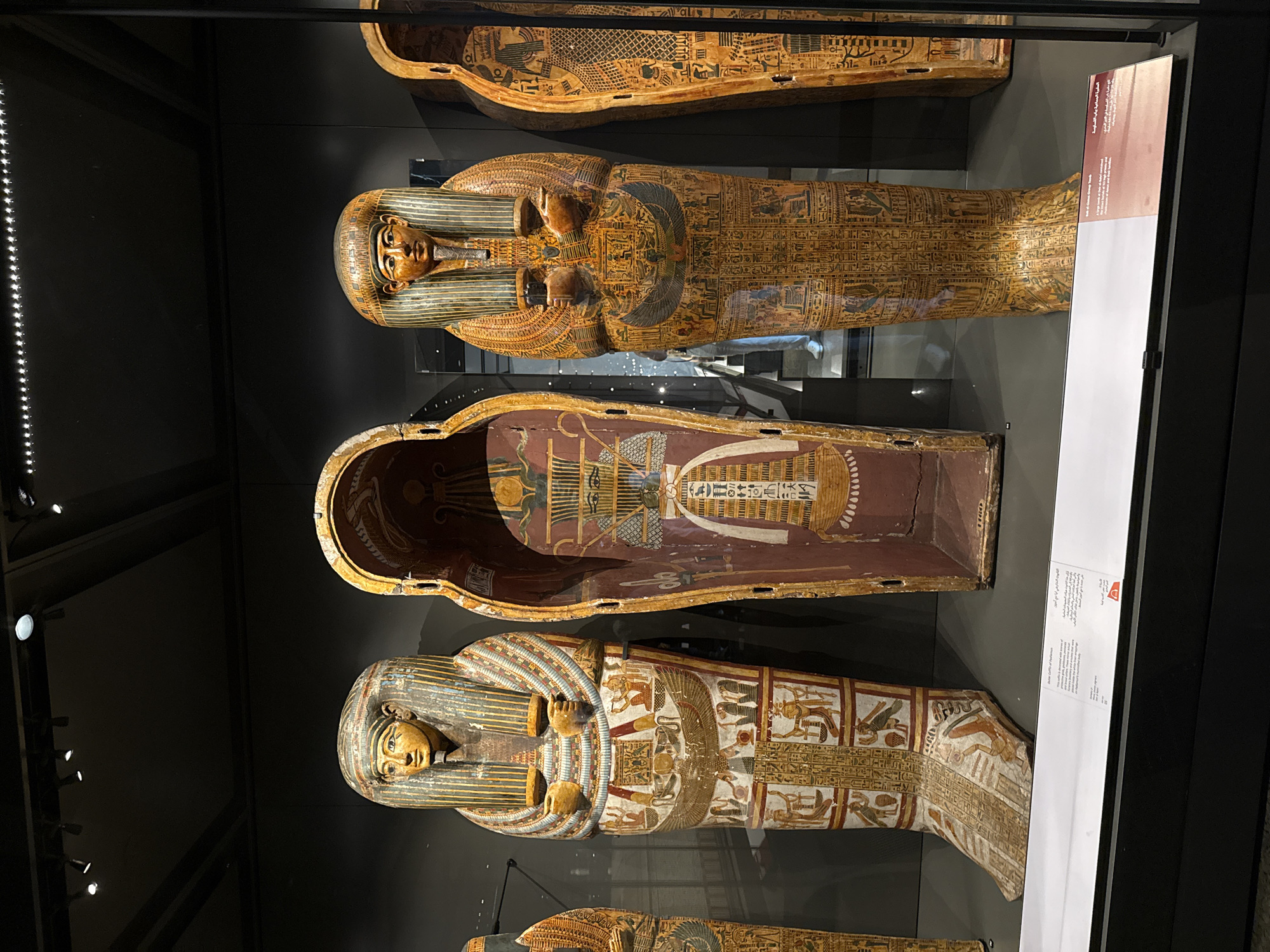 The image shows three ancient Egyptian sarcophagi displayed in a museum exhibit. The sarcophagi are elaborately decorated with hieroglyphs and colorful imagery. The sarcophagus in the center is open, revealing the interior with painted scenes. The other two sarcophagi are closed and stand upright. The lighting in the exhibit highlights the details of the artifacts. There is a label in English and Arabic at the bottom of the display. The environment appears to be a dimly lit museum hall with a dark background. The city of Kafrat Nassar is not depicted in this image.