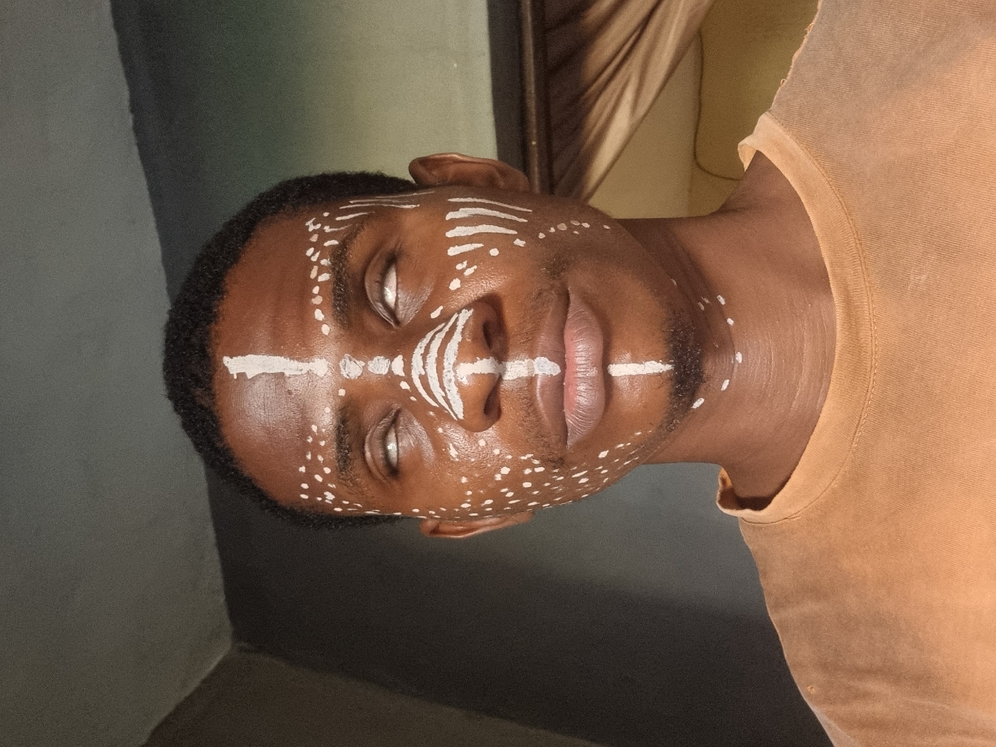 A person is depicted in the image. The individual has brown skin and short black hair. White paint is applied to their face in various patterns including a horizontal line across the forehead, lines down the nose, and dots on the cheeks. The person is wearing a light orange t-shirt. The background is a combination of gray and beige tones, with a textured surface. The location and city are unknown.