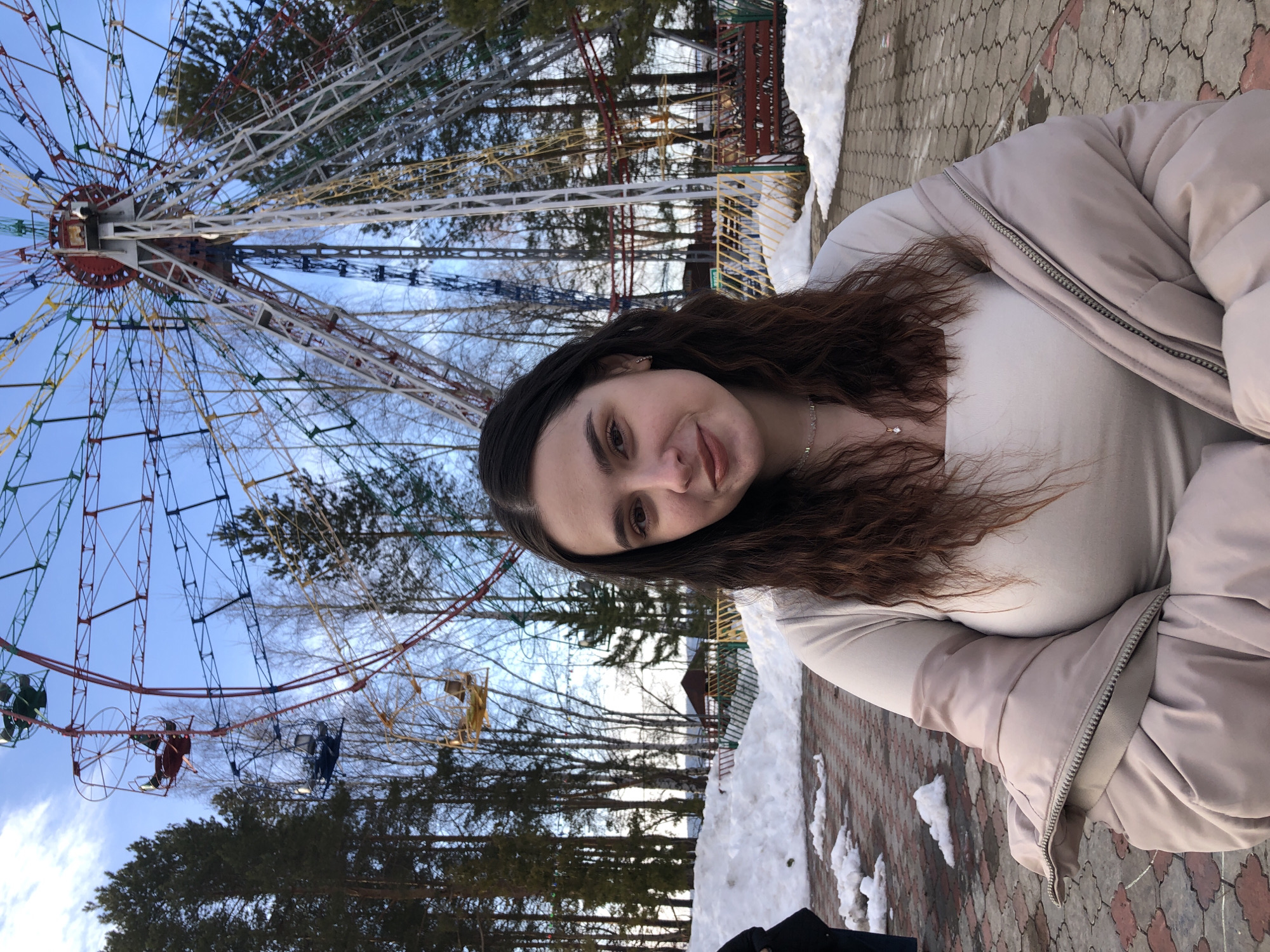 A young adult female is positioned in the foreground of an outdoor park setting within the Park Kultury i Otdykha (Park of Culture and Rest) in Chaikovsky, Perm Krai, Russia. She has dark, wavy hair, dark eyes, and fair skin, with a faint smile directed towards the viewer. She is wearing a light beige square-necked top and a light pink/beige puffer jacket draped unzipped over her shoulders. A delicate necklace with a small clear pendant is visible.
In the background, a large, multicolored Ferris wheel dominates the upper frame, featuring red, blue, yellow, white, and green structural elements with multiple passenger gondolas in matching colors. Some gondolas appear empty. Tall evergreen and bare deciduous trees frame the amusement ride and extend across the mid-ground. The ground where the woman stands is paved with reddish-brown interlocking pavers, surrounded by melting snow patches. A small, dark-roofed wooden building is partially visible in the distant background to the left. The sky above is clear blue with scattered white clouds. A partial figure in dark clothing is visible on the far left edge of the frame.