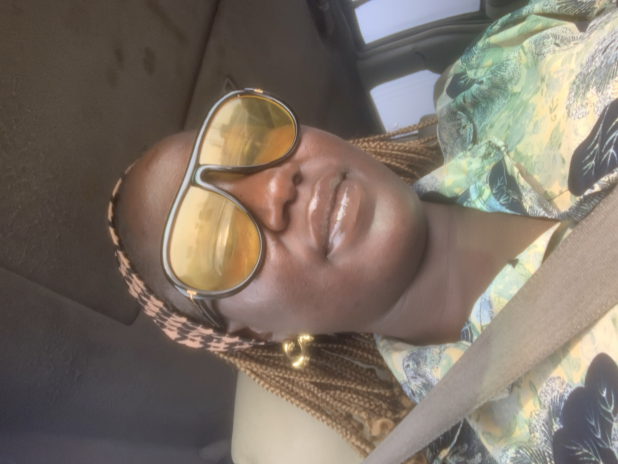 A woman with dark skin and light brown box braids is seated inside a vehicle, likely in Elelenwo, Nigeria. She is wearing a patterned headband, a button-up shirt with a green, yellow, and black abstract or tropical print, and a light brown seatbelt across her chest. Large, dark-framed aviator-style sunglasses with reflective yellow lenses obscure her eyes; reflections in the lenses show an outdoor environment with structures and light. A glossy substance is applied to her lips, and a large, gold-toned, C-shaped earring is visible on her left ear. The vehicle's headliner, appearing dark grey, is visible above her, and a car window is visible to her right, showing a hazy bright sky or distant view.