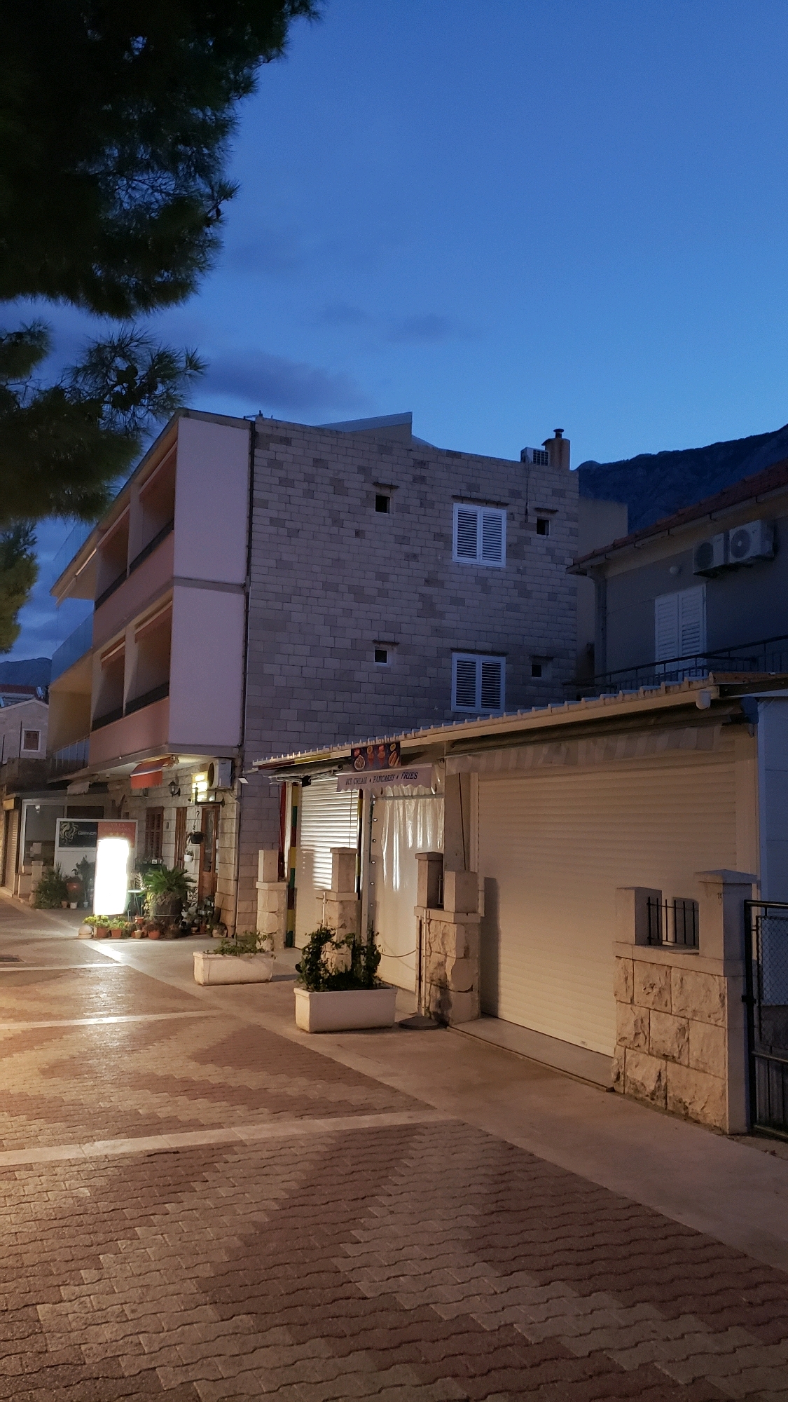 This is a vertical, medium-shot, outdoor, nighttime photograph of a street scene in Općina Tučepi, Croatia. The sky is a deep, clear blue with a few dark clouds. On the left, a large pine tree branch with dark green needles extends into the frame from the top left. In the center and right, a three-story building with a light-colored brick facade and balconies is visible. The building has white-shuttered windows and air conditioning units mounted on the exterior.
In front of the three-story building, a lower, single-story structure with a white roller shutter door is present. Above this shutter, a sign reads "ICE CREAM • PANCAKES • FRIES". Stone pillars with decorative ironwork are visible in front of this structure. To the right of this, a darker building with a red-tiled roof is partially visible, also with white-shuttered windows and an air conditioning unit.
The foreground is dominated by a paved walkway constructed of reddish-brown interlocking pavers arranged in a staggered pattern. Two large, rectangular white planters containing greenery are positioned on the walkway. The lighting in the scene appears to be a mix of streetlights and interior lights from the buildings, casting warm tones on the pavement and facades. The overall atmosphere is quiet and somewhat serene, suggesting a resort town at dusk or after dark. There are no people or animals visible in the frame.