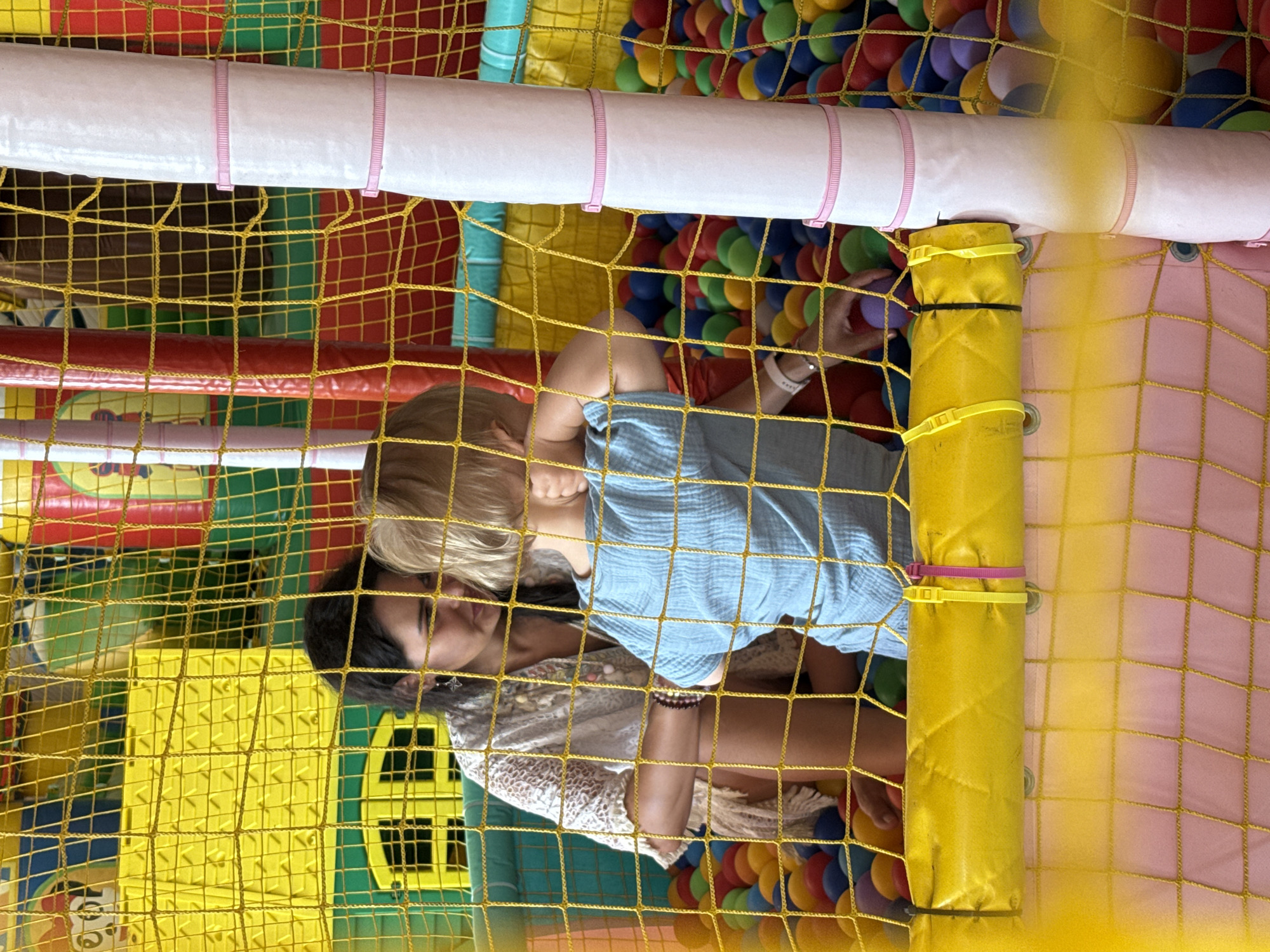 A woman and a child are in a ball pit inside a brightly colored indoor playground. The woman, with dark hair, is wearing a light-colored, patterned open-weave top and is leaning towards the child. The child, with blonde hair, is wearing a light blue t-shirt and has their back to the camera, with one hand covering their face. The ball pit is filled with numerous brightly colored balls, including red, blue, green, and yellow. The playground structure behind them features yellow, green, and red panels. The scene is viewed through a yellow mesh net, with a white padded pillar with pink bands visible on the right side. The woman is holding a ball. The background suggests an indoor recreational facility.