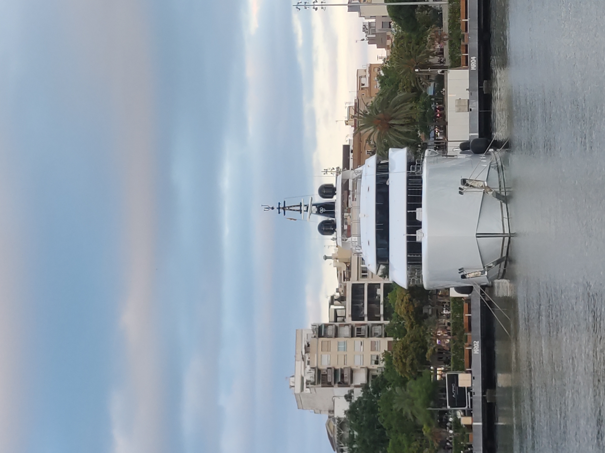 The image shows a large white yacht docked in a harbor. In the background, there are several multi-story buildings with balconies, palm trees, and other greenery. The sky is overcast with clouds. The yacht has a sleek, modern design with multiple levels and a prominent mast. A gangway extends from the yacht to the dock, and mooring lines are visible. The dock has some markings, including "MR02" and "MR04". There are a few people visible on the dock in the background. The water in the foreground appears calm. The scene suggests a waterfront area in a coastal city, likely during daylight hours. This appears to be the Port Olímpic in Barcelona, Spain, given the architectural style of the buildings and the presence of such large yachts.