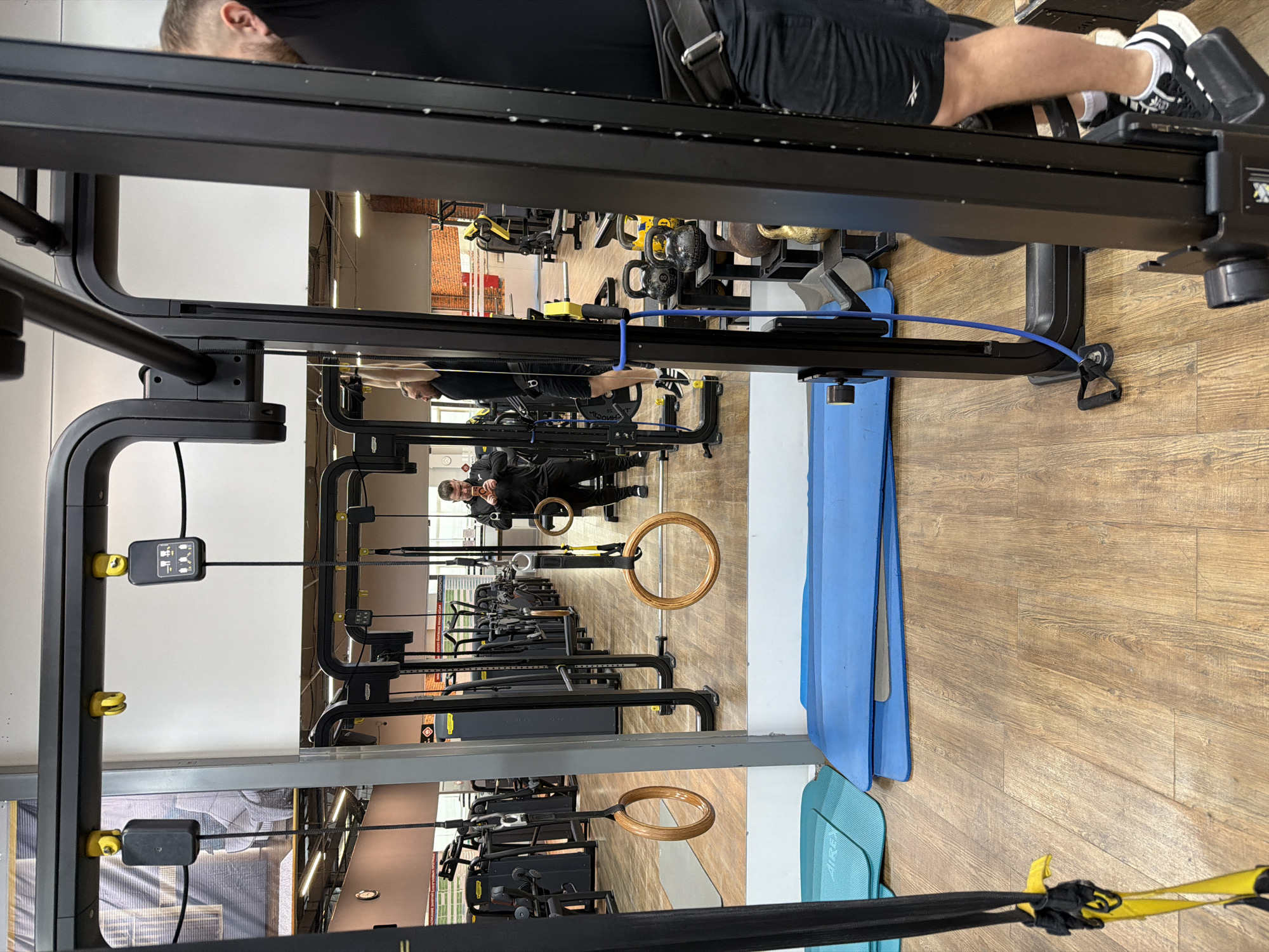 The image depicts the interior of a gym, likely in Chaikovsky, Russia, based on the prompt. The gym contains various weight machines, including leg press machines and cable pulley systems. Several sets of gymnastic rings are also visible, hanging from the ceiling.
A man is visible in the background, reflected in a mirror. He appears to be taking a photo of himself using a smartphone. Another man is on a rowing machine in the top portion of the image.
The gym floor is wooden, and blue and green exercise mats are stacked against a wall. There is gym equipment such as kettle bells visible.