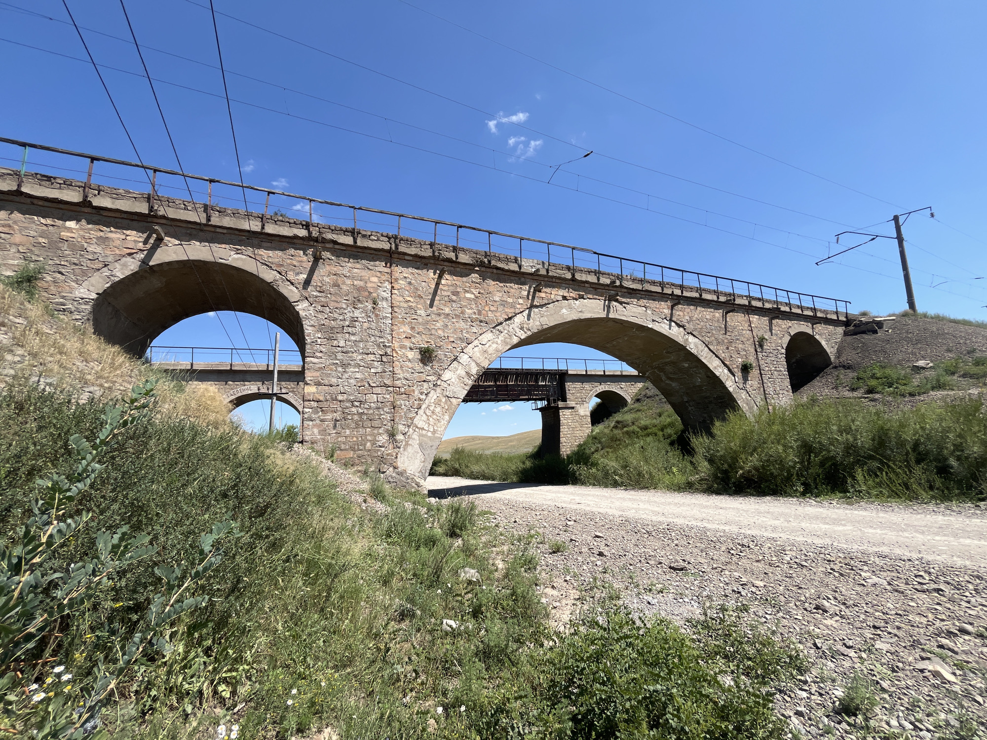 The image shows a dry, dusty rural road passing under a large, old stone arched railway bridge. The bridge is made of rough-hewn stones and appears to be in disrepair in some areas, with visible concrete patches and rusty metal railings. The road is unpaved and covered with gravel and dirt. On either side of the road, there is dry grass and green bushes, suggesting a semi-arid environment. In the background, rolling hills are visible under a clear blue sky. Another, smaller bridge or viaduct can be seen further in the distance, also made of stone arches and supporting what appears to be a railway track with overhead electrical lines. The context provided places the scene in Astakhovka, Kazakhstan. There are no people or animals visible in the image. The overall impression is one of a remote, somewhat neglected infrastructure in a natural, open landscape.