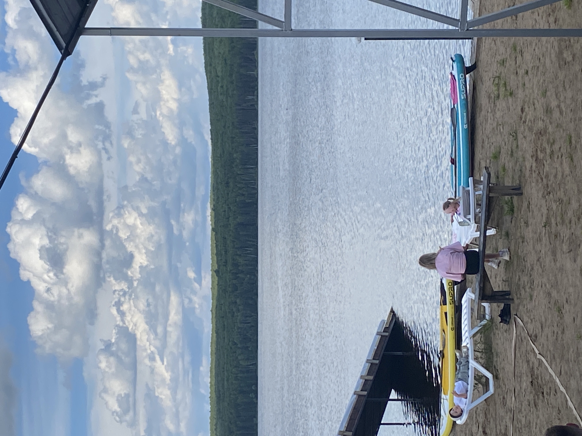 In Chaikovsky, Russia, on a sandy beach beside a wide, calm body of water, two stand-up paddleboards are resting on the shore. One is yellow and the other is blue with pink accents, bearing the "SUPRO" logo. In the foreground, a person lies on a white lounge chair, appearing to be relaxing or sunbathing. Nearby, a woman sits on a wooden bench, facing away from the camera. Behind them, a wooden dock extends into the water. The background features a dense forest lining the opposite shore, with a cloudy sky overhead. The overall scene depicts a tranquil day at the waterfront.