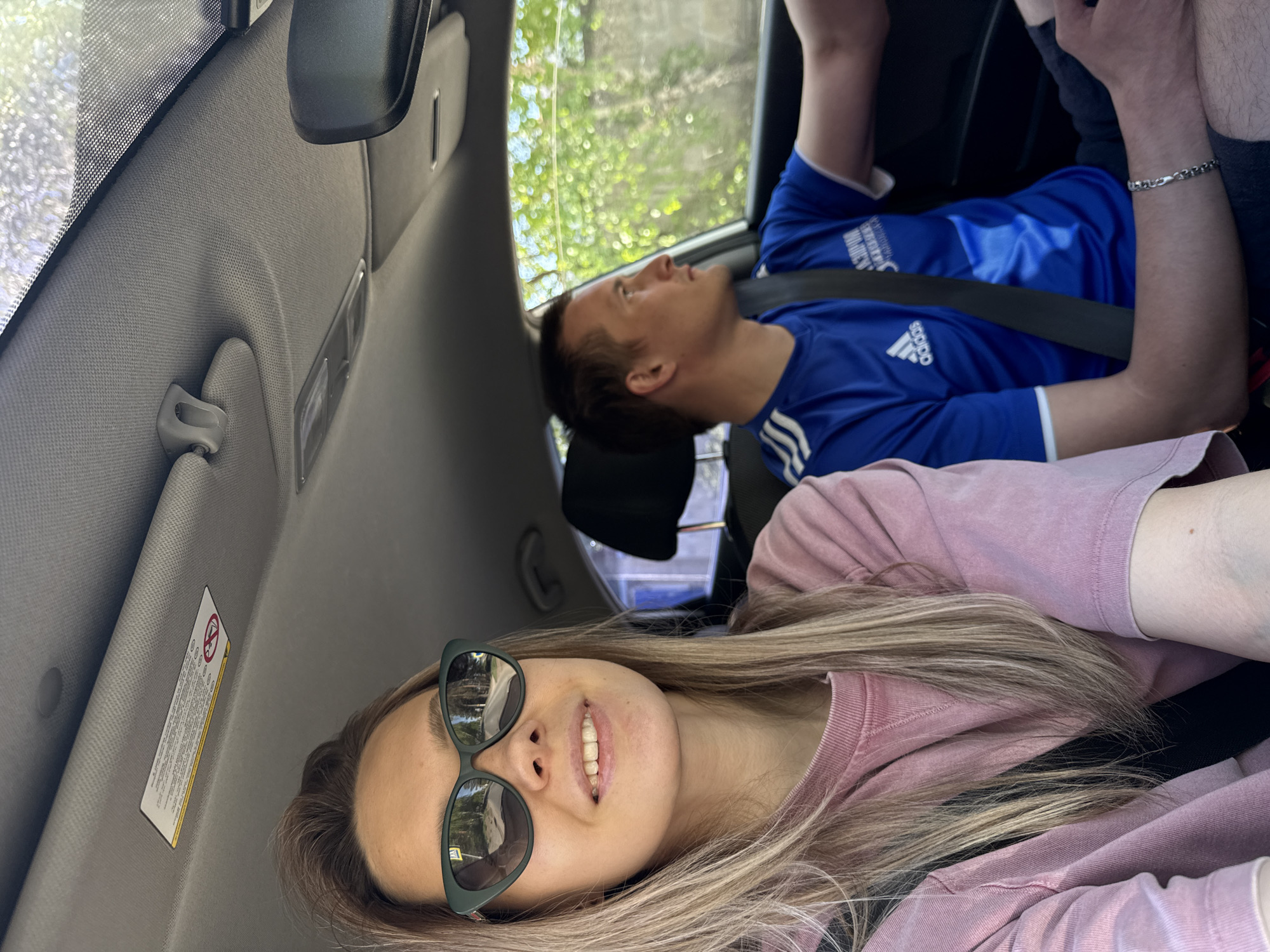 The image shows two people inside a car. The person in the foreground is a woman wearing sunglasses and a pink sweatshirt, smiling at the camera. Behind her is a man in a blue Adidas shirt, looking upwards. Both are wearing seatbelts. The interior of the car is visible, including the dashboard, sun visor, and windows. The location of the scene is not discernible as Noviy, Russia.