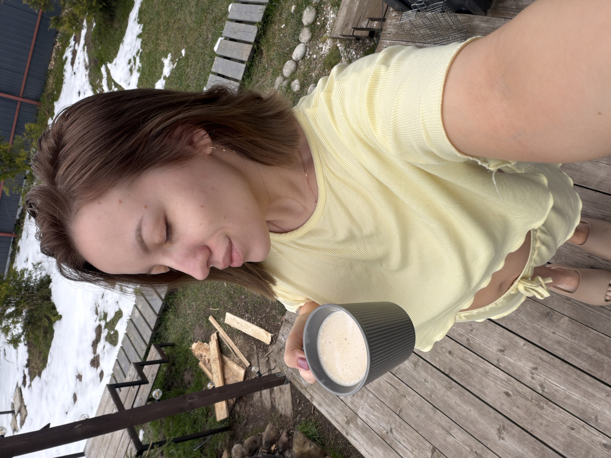 A woman in a yellow top and sandals is standing on a wooden deck, holding a dark gray mug filled with a light-colored beverage. The location appears to be outdoors, likely in a backyard or garden. There is snow and grass visible in the background, along with a wooden walkway and some logs or firewood. The perspective is a high-angle selfie. It is unlikely that the location is in the city of Бунятино, Russia.