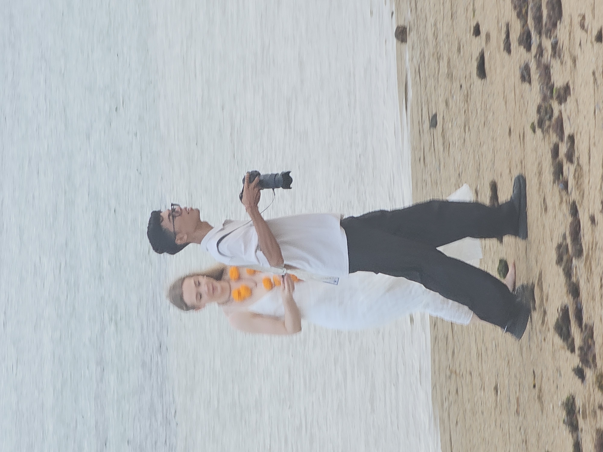 Two individuals, a woman and a man, are walking on a sandy beach near the water's edge. The woman is on the left, wearing a white dress and an orange flower garland around her neck. She has blonde hair. The man is on the right, wearing a white short-sleeved shirt, dark pants, and glasses. He is holding a camera and appears to be taking photos. The background consists of the ocean. The scene is likely in a coastal area of Denpasar, Indonesia, known for its beaches.
