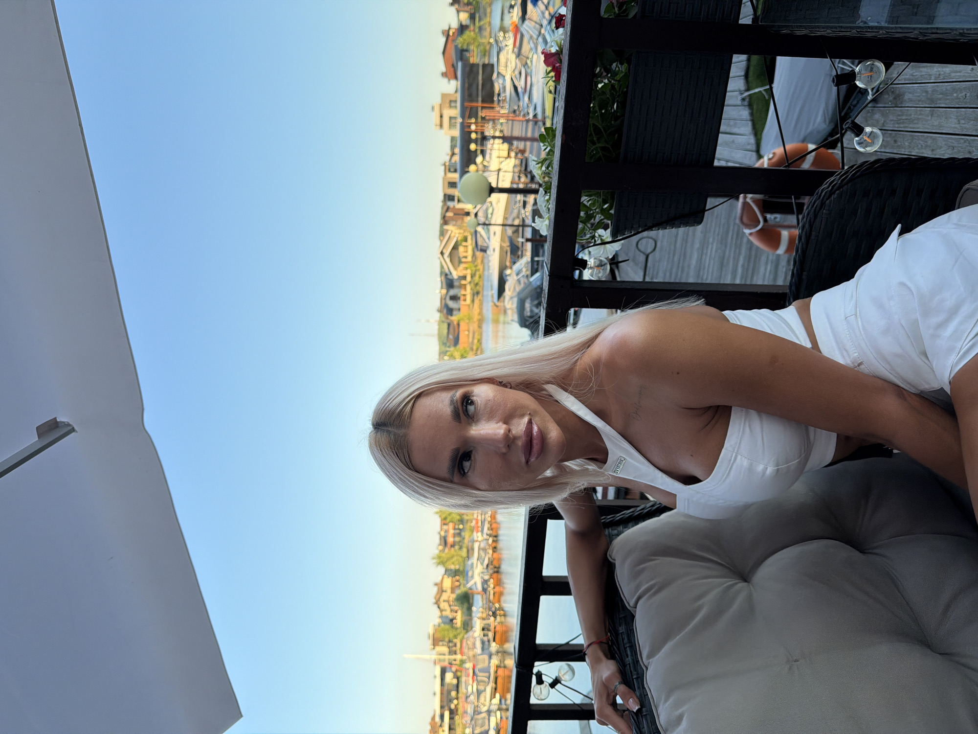 In Kazan, Russia, a woman with blonde hair is sitting on a chair outdoors, looking to her right. She is wearing a white top and shorts. Behind her, a body of water is visible, with numerous boats docked. The scene appears to be a marina or waterfront area. The sky is clear and blue, suggesting it is daytime.
