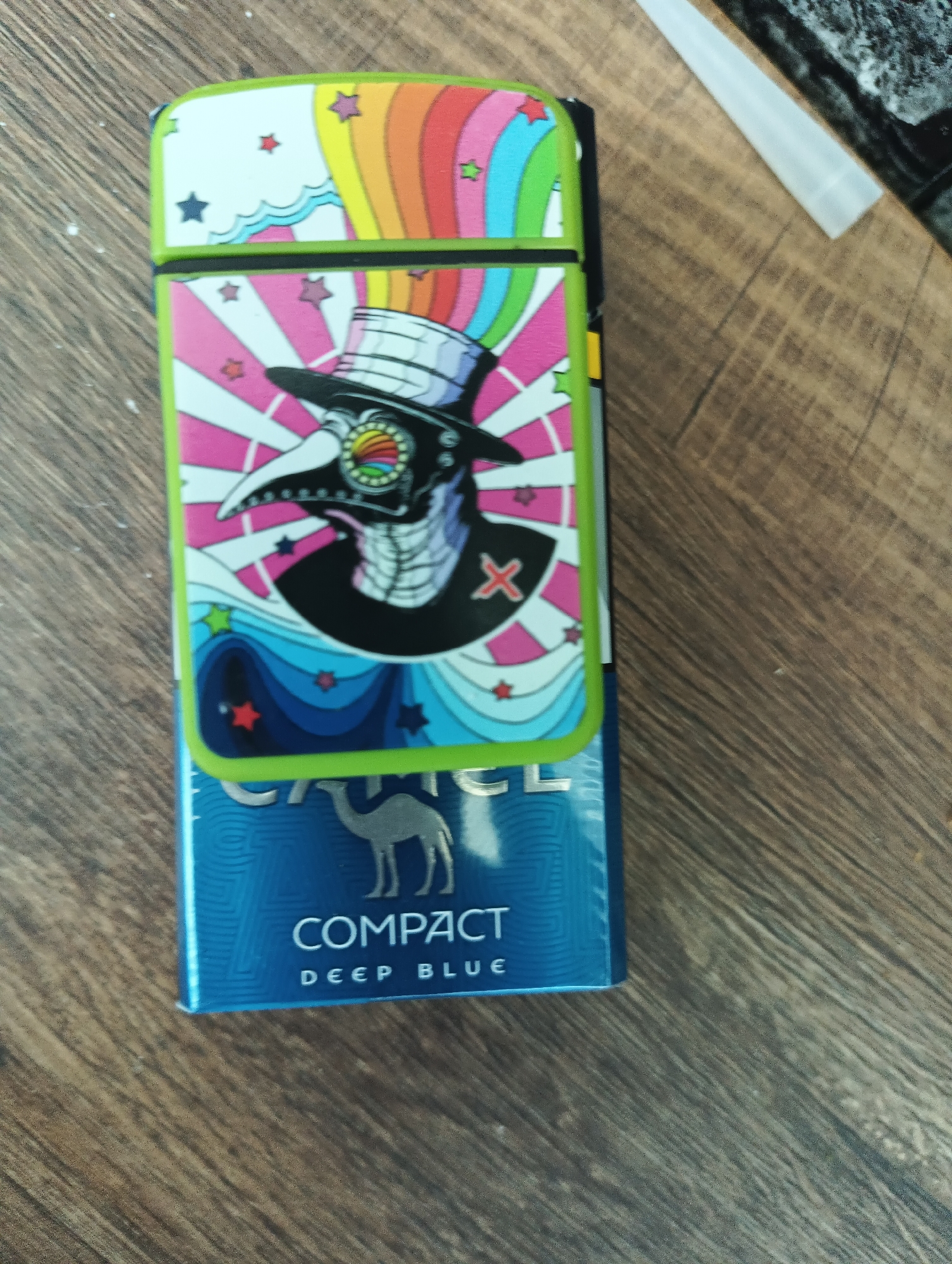 A close-up, high-angle shot shows a pack of Camel cigarettes, specifically "Compact Deep Blue," resting on a wooden surface. The cigarette pack is partially covered by a lighter with a vibrant, psychedelic design. The lighter features a rainbow arch at the top, with colorful stars and a stylized, plague-doctor-like figure in the center. The figure wears a hat and a mask with a rainbow-colored eye detail. The background is a wooden table, with some white dust specks visible. There is no discernible location in Karnobat, Bulgaria.