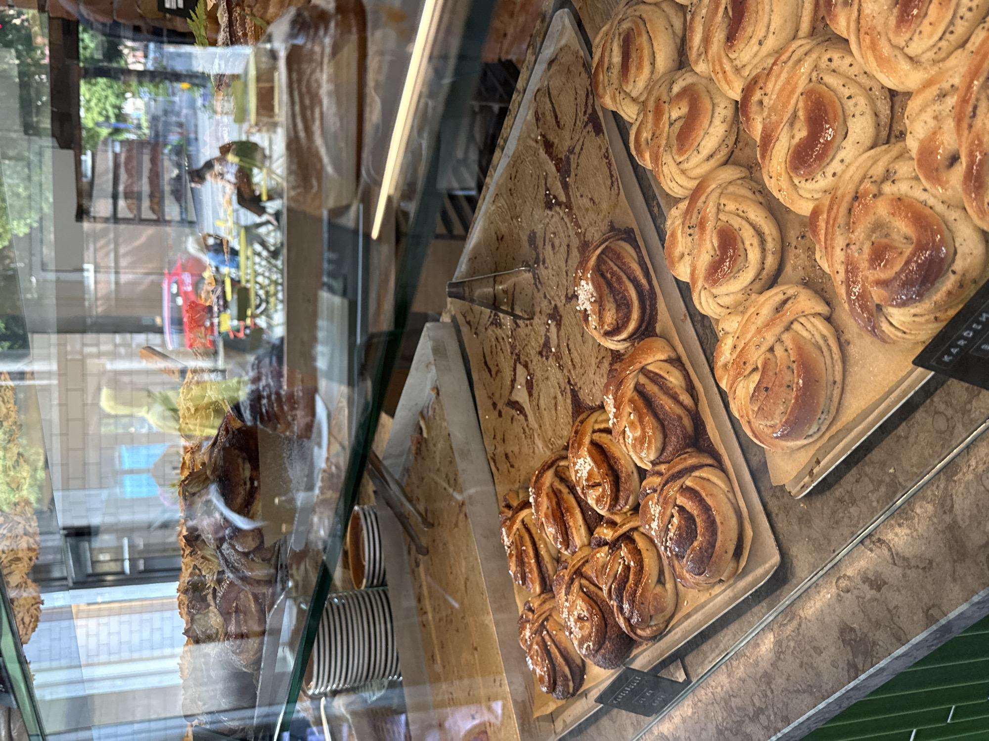 The image shows a display case filled with baked goods, likely from a bakery or cafe in Stockholm, Sweden. The baked goods are arranged on trays and appear to be sweet pastries, with some featuring a swirl pattern and a dusting of sugar or spice. In the background, visible through the glass of the display case, are reflections of people and a street scene, including tables and chairs outside, a red vehicle, and trees. The overall impression is of a well-stocked bakery, with a focus on visually appealing pastries.