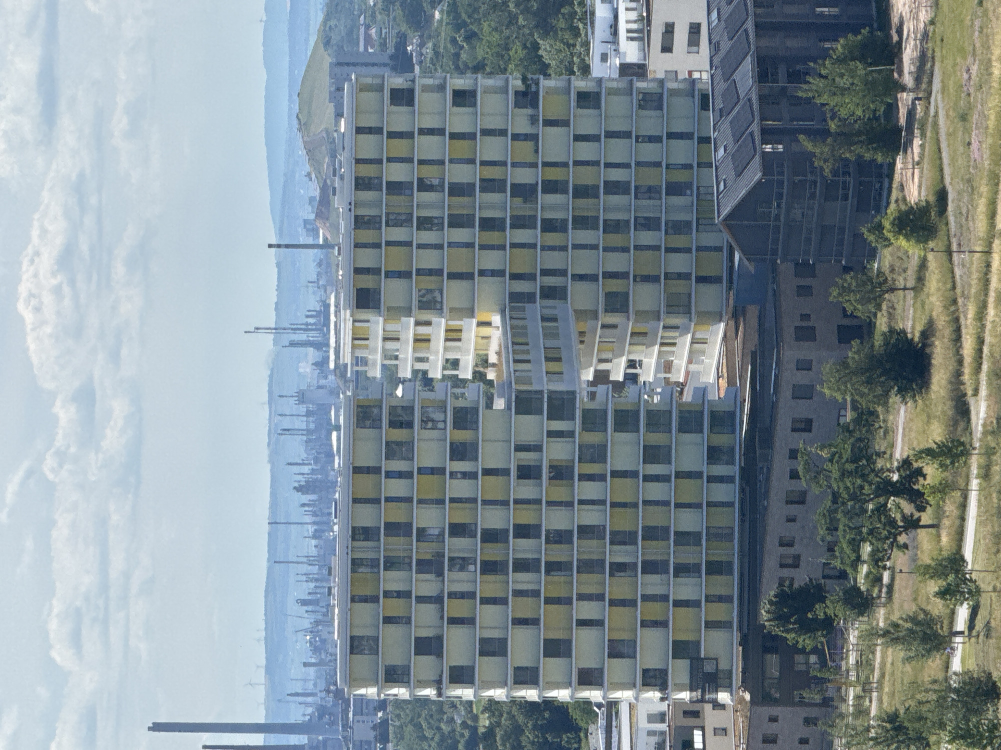 The image shows an aerial view of two modern, multi-story residential buildings with a facade of light yellow and light blue panels and dark window frames. The buildings are situated in a cityscape, with a lower, darker building with a sloped roof and solar panels visible in the foreground. Beyond these buildings, there is a background of trees, and further in the distance, an industrial area with numerous tall chimneys and factory structures. The sky is partly cloudy with a pale blue hue. The scene is set in Mannheim, Germany, specifically in the area of the former Rheinau-Kaserne, which has been redeveloped into a residential district. The overall impression is one of urban development integrating modern architecture with existing industrial landscapes. No people are clearly visible in the image.