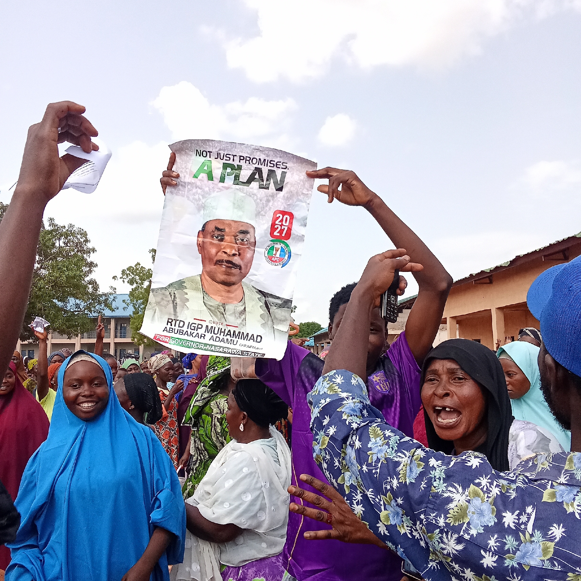 A crowd of people in Akwanga, Nigeria are gathered, likely at a political rally or demonstration. They are holding up campaign posters for RTD IGP Muhammad Abubakar Adamu for Governor, Nasarawa State, with the slogan "Not Just Promises. A Plan 2027." Many are wearing colorful traditional clothing, and some are smiling and cheering, suggesting their support for the candidate. One person is holding up what appears to be campaign literature, while another person is recording the event with an older model cell phone. The environment consists of buildings and trees in the background, partially obscured by the crowd. The sky overhead is mostly white and clouded.