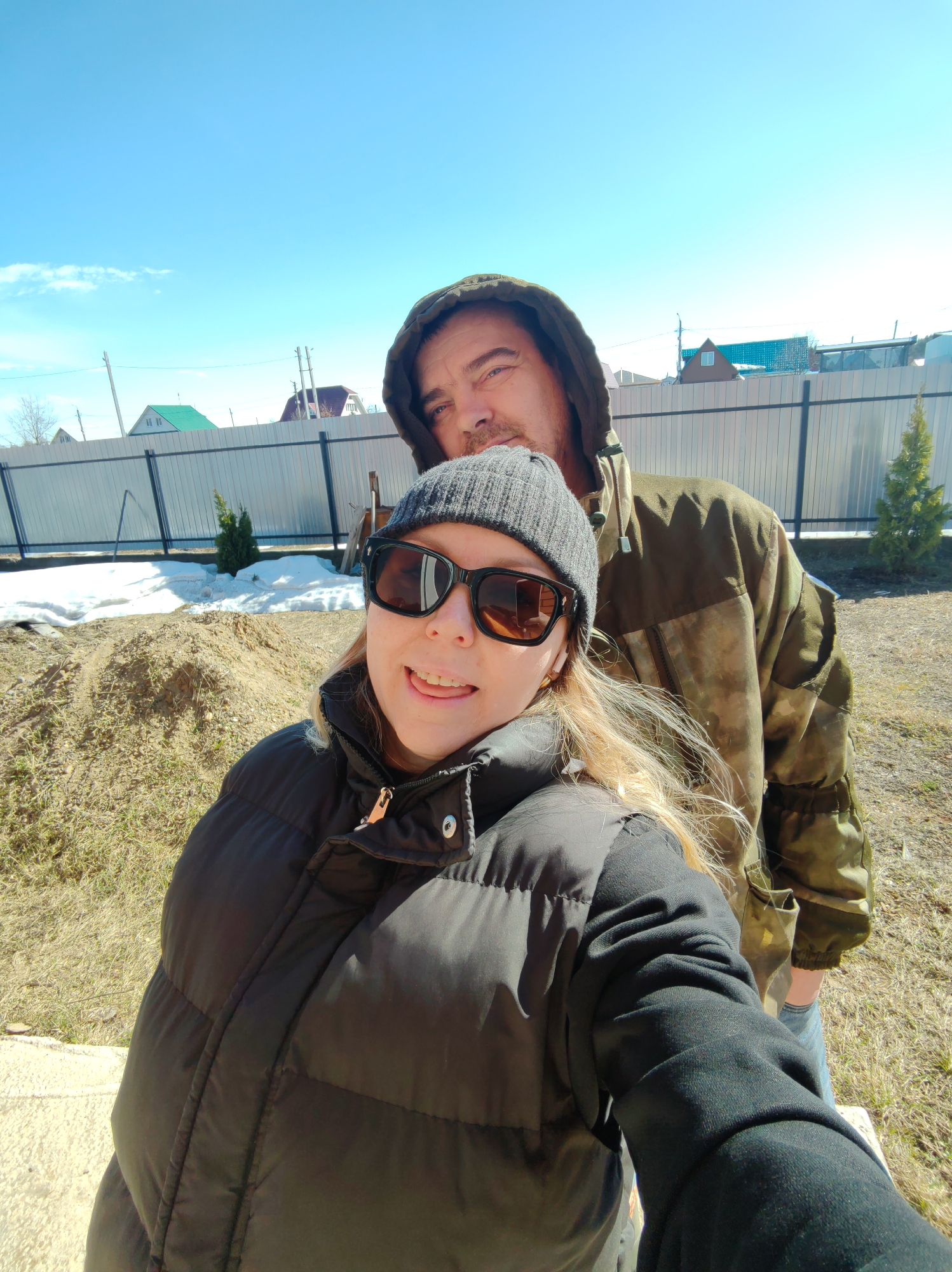 A selfie of two individuals is taken outdoors in what appears to be a residential area. A woman, wearing a black padded jacket, a grey knit hat, and dark sunglasses, is holding the camera, her tongue slightly visible. Behind her is a man with a hood up, wearing a camouflage-patterned jacket. In the background, there is a light-colored fence, a pile of dirt, some remaining patches of snow, and a few small evergreen trees. Further back, houses with varied roof colors can be seen under a blue sky with scattered clouds. Given the presence of snow remnants and the style of housing, the location is likely in a colder climate, possibly Chaikovsky, Russia, as indicated. The action depicted is a selfie, capturing the two people in a casual setting outdoors.