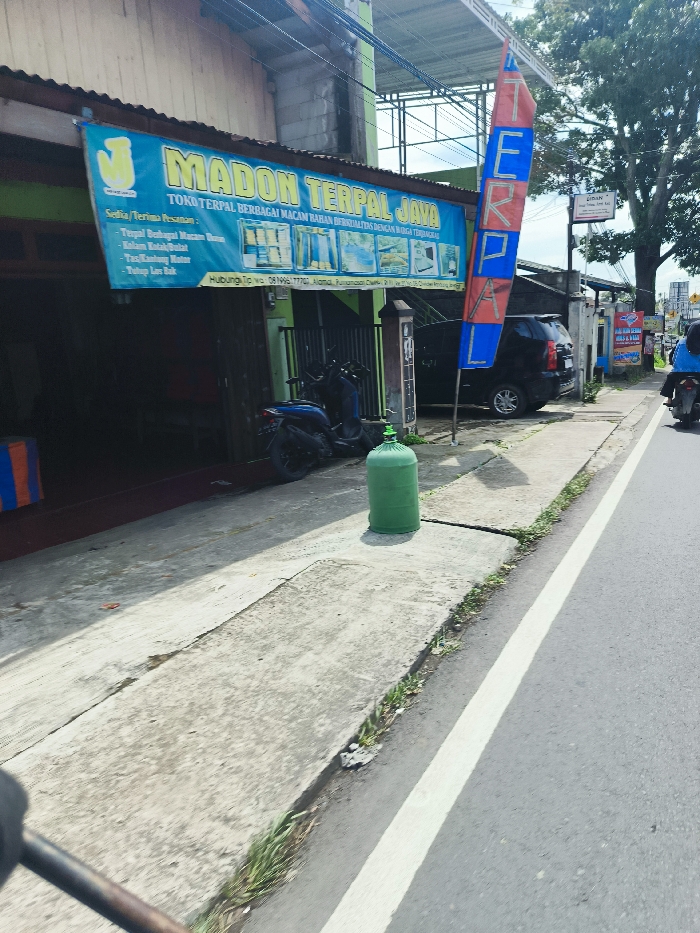 The image shows a street in Ciwidey, Indonesia, a town located at coordinates -7.106 latitude, 107.457 longitude. There is a large blue and yellow sign on the front of a shop advertising "Madon Terpal Java," a store selling a variety of goods. A blue and red flag with the word "TERPAL" on it stands in front of the shop. A black car is parked on the street. There is a green gas cylinder sitting on the sidewalk in front of the shop. A motorbike is parked in front of the shop. The road is paved, with a white line separating it from the sidewalk. The sidewalk is lined with grass and trees, including a large tree with a canopy of leaves. The sun is shining, and there are no people visible in the image. There is a building in the background, as well as other signage. The image is taken from the perspective of someone walking on the sidewalk.