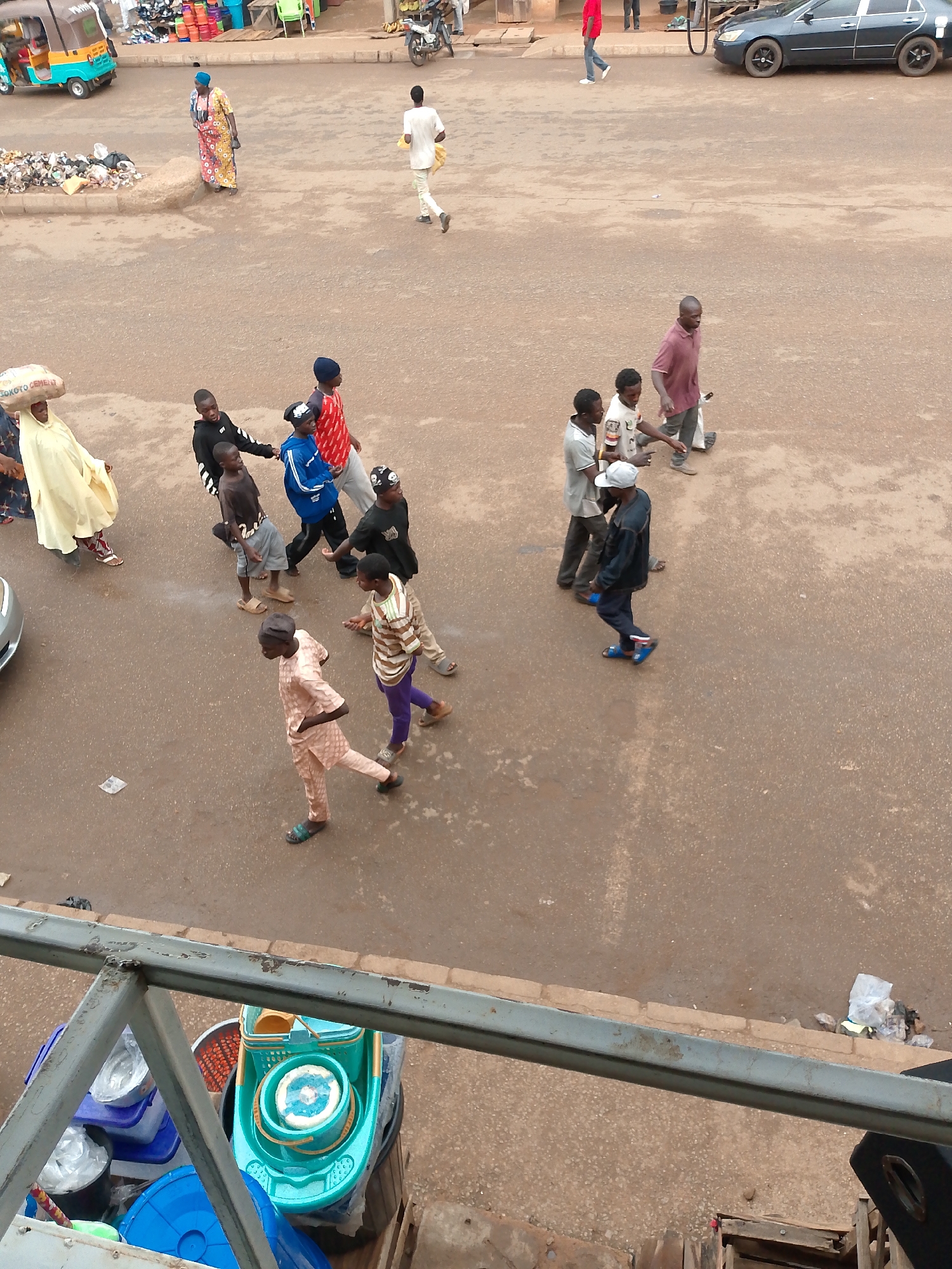 The image is taken from an elevated position in Bukuru, Nigeria, overlooking a street scene. Several people are walking on the street. A woman carries a sack of "SOKOTO CEMENT" on her head, a group of children walk closely together with some holding onto one another. Further along the street, three men walk in the same direction. A man in a white shirt walks away from the main group of people. Parked vehicles, including a car and a tricycle "KEKE", are visible in the background. In the lower part of the frame, an object with blue and green cleaning materials is partially visible behind a metal railing.