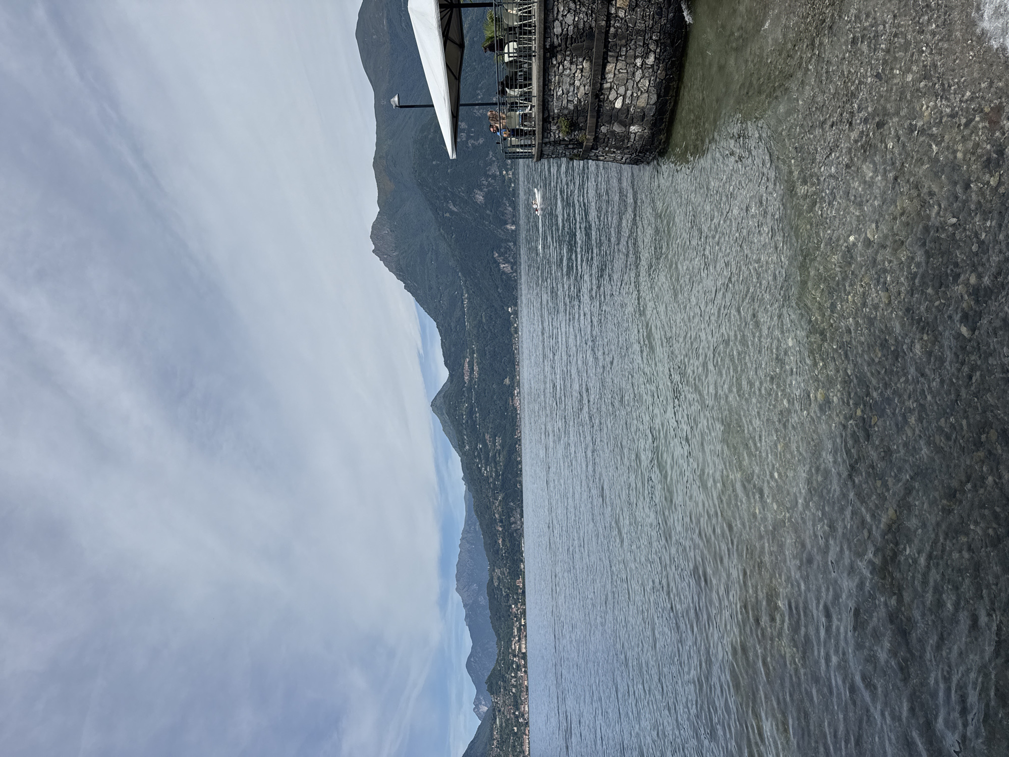 The image depicts a serene lakeside scene in Gittana, Italy. In the foreground, clear water laps against a pebble-strewn shore. The mid-ground is dominated by a calm lake, reflecting the overcast sky. In the background, a range of lush green mountains rise, dotted with small villages. On the right side of the frame, a stone-built structure juts out into the water, topped by a patio with tables and chairs. A white umbrella provides shade for a few patrons enjoying the view. A small boat is visible in the distance on the lake. The overall atmosphere is peaceful and picturesque.