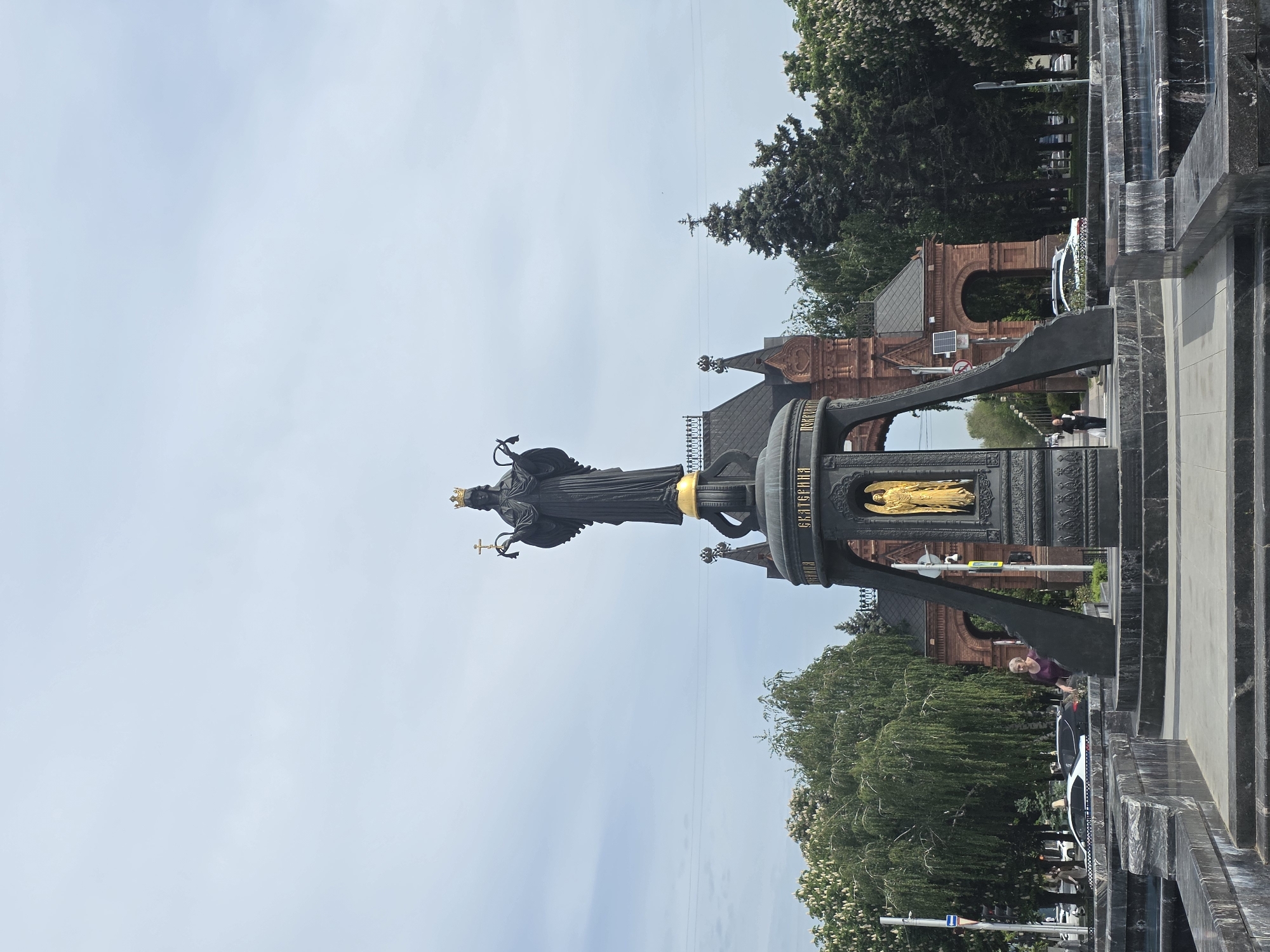 The image shows a monument to Catherine the Great in Krasnodar, Russia. The statue is bronze and depicts Catherine holding a cross and crown, with an angel encased in gold below her. In the background is an ornate brick gate. A woman in a purple shirt stands by the monument, and cars and people can be seen along the sidewalk. The sky is light blue.