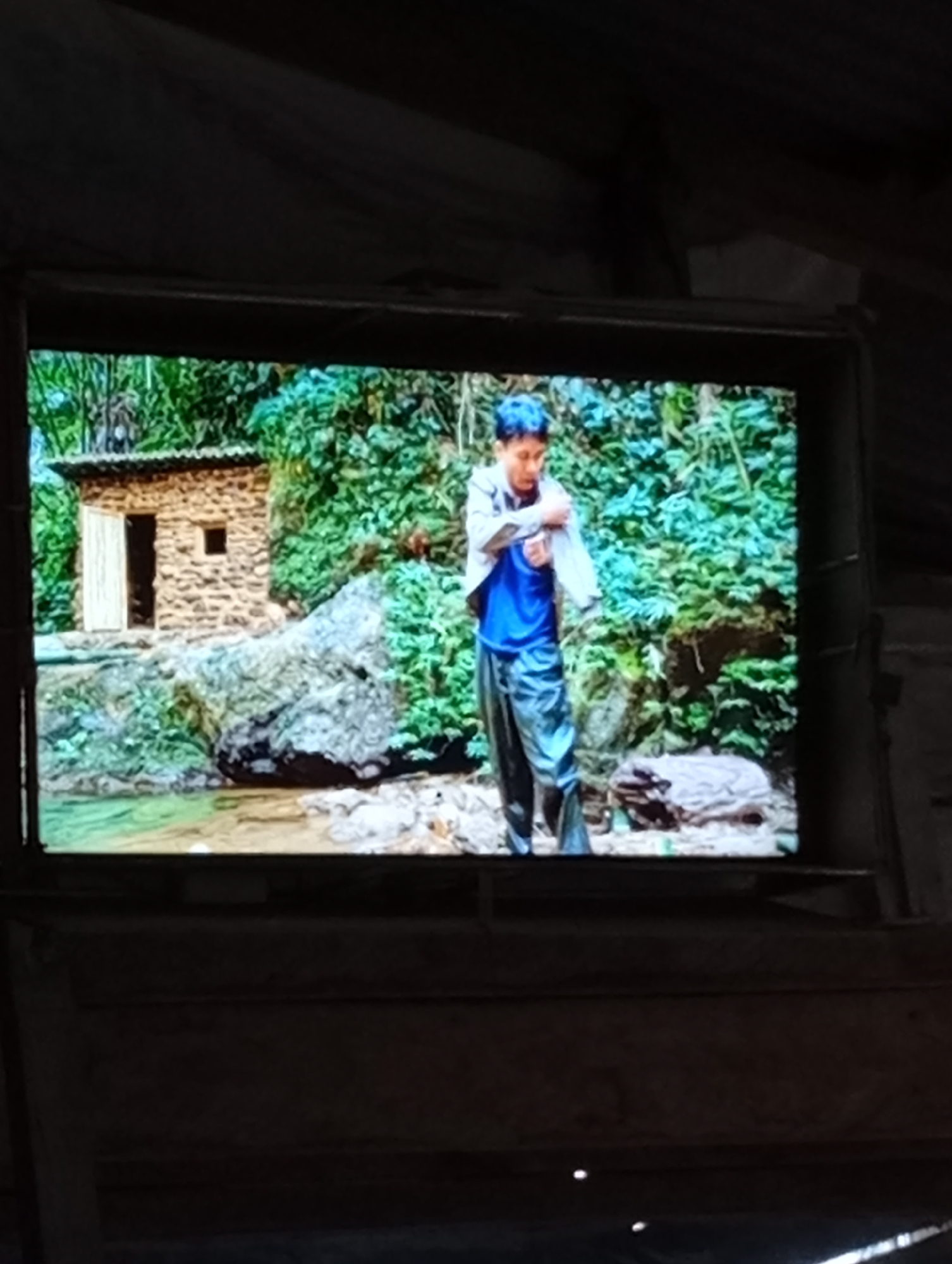 The image shows a television screen displaying a scene of a young person standing in a natural environment. The person is wearing a blue shirt and dark pants. Behind them, there is lush green foliage and a small, stone building with an open door. A body of water, possibly a stream or river, is visible in the foreground, with rocks surrounding it. The television screen is dark-framed and appears to be old. The surrounding environment in the image of the room where the television is located is dimly lit and appears to be rustic, with wooden structures visible. The location of Lhokseumawe, Indonesia is not visually identifiable within the provided image content.