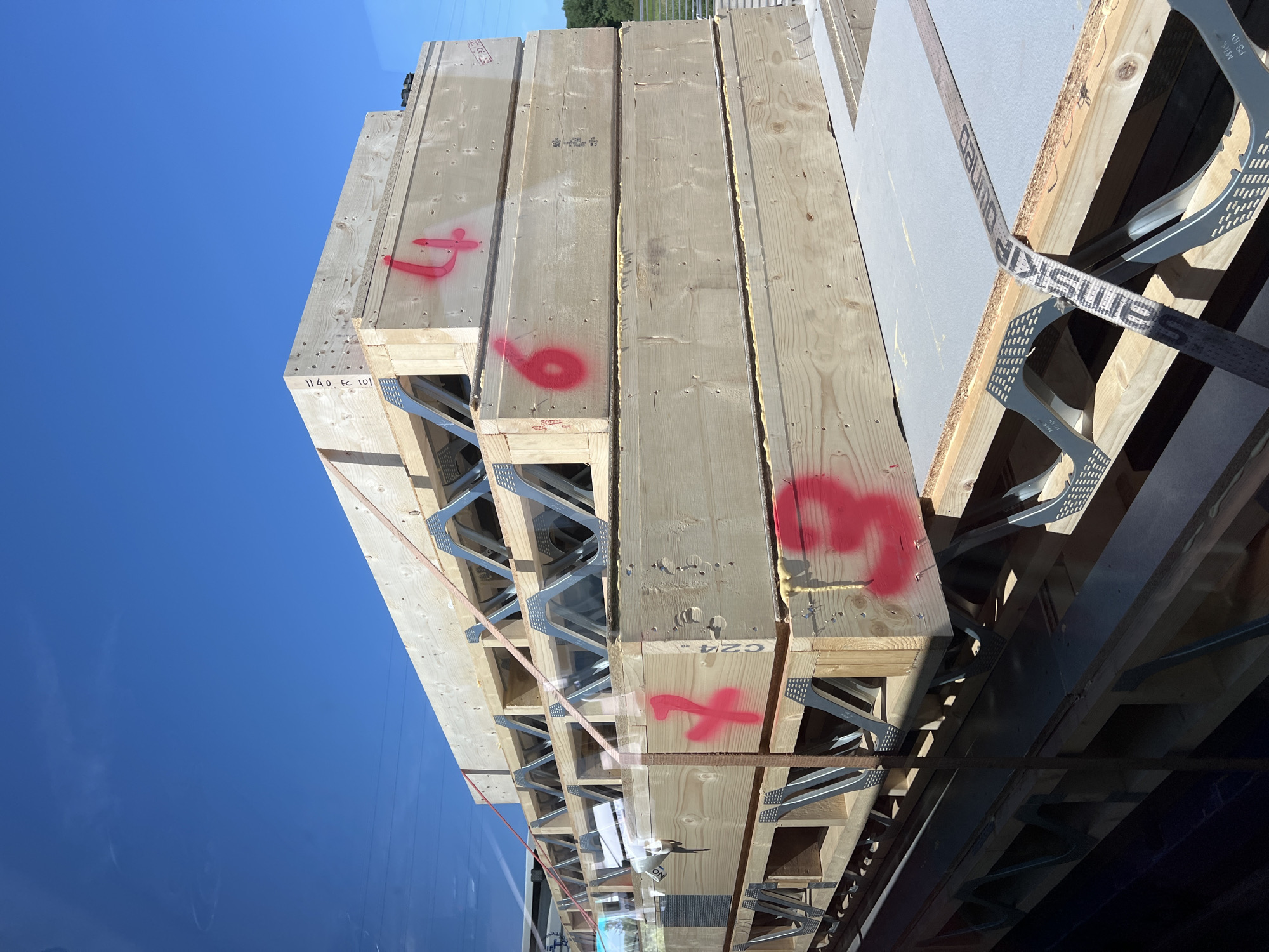 The image shows a stack of large wooden beams, possibly for construction, secured with straps and metal bracing. Some of the beams are marked with red spray-painted numbers, including "4", "6", and "3". The beams are arranged in a tiered fashion, with metal brackets supporting them. The scene appears to be outdoors under a clear blue sky, with hints of distant trees and possibly power lines visible in the background. The perspective suggests the photo may have been taken from a slightly elevated position, perhaps from another vehicle or structure. The image does not provide any specific visual cues that would allow for the determination of its location in Letterkenny, Ireland.