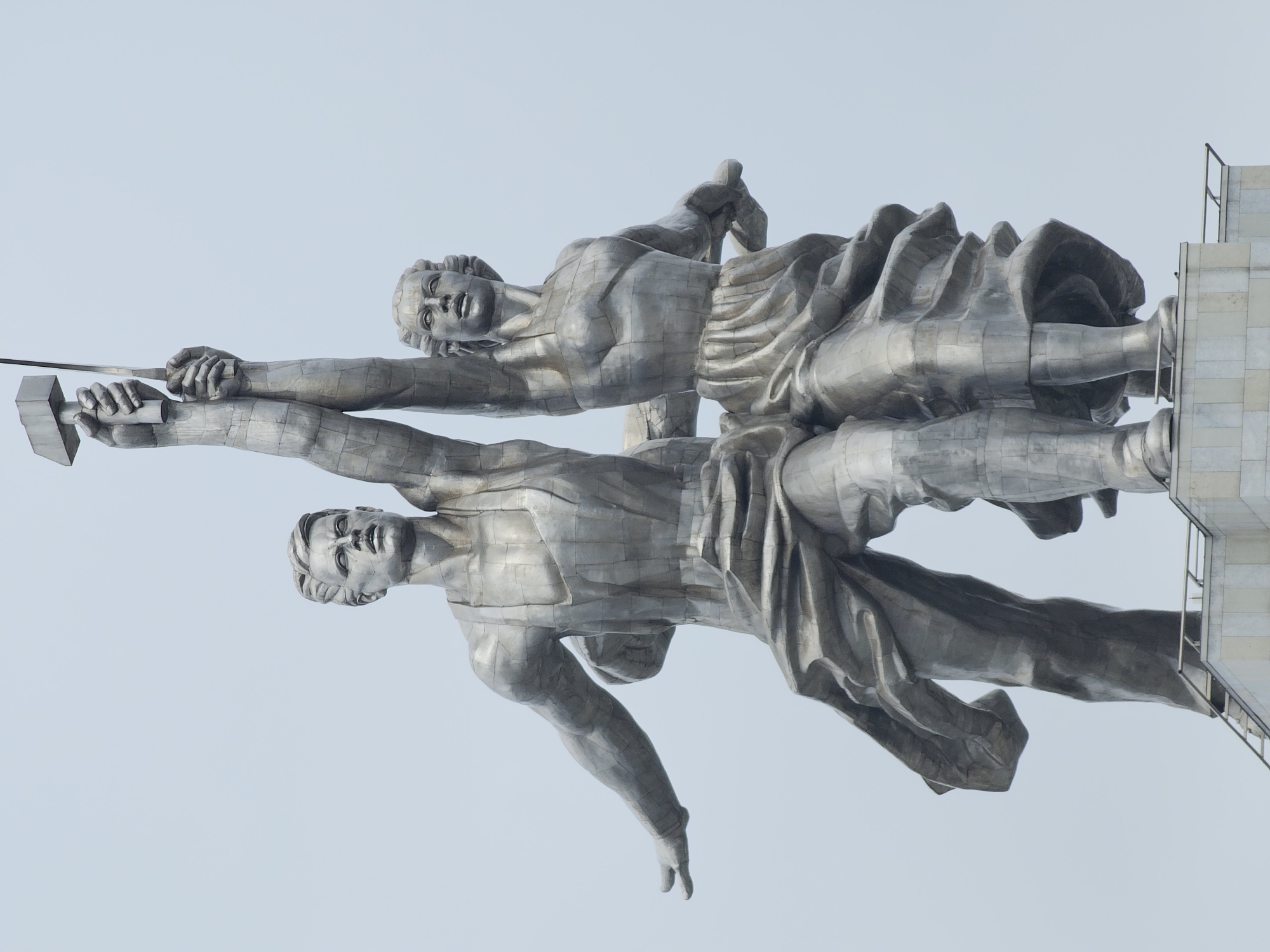 This image presents an upward-looking view of the monumental "Worker and Kolkhoz Woman" sculpture, located in Moscow, Russia. The main subjects are two colossal, stylized human figures, one male and one female, crafted from metallic panels with visible seams that create a distinctive, segmented appearance. The figures are depicted in a powerful, unified motion, striding forward and upward.
The male figure, positioned lower and slightly in front, holds a hammer aloft in his right hand. His face is resolute, gazing forward and upward. The female figure, positioned behind and slightly above, holds a sickle in her left hand, which is raised in conjunction with the hammer, forming the iconic hammer and sickle symbol. Her expression also conveys determination and aspiration. Both figures wear stylized clothing, with drapery suggesting movement and energy.
The sculpture is set against a clear, light blue sky, indicating bright daytime conditions and fair weather. On the far right, a tall, grey, rectilinear structure, likely part of the monument's pedestal or supporting architecture, is visible, providing a contextual anchor for the soaring figures. The overall impression is one of immense strength, unity, and forward-looking optimism, symbolizing the combined power of the working class and peasantry. No text is visible in the image.