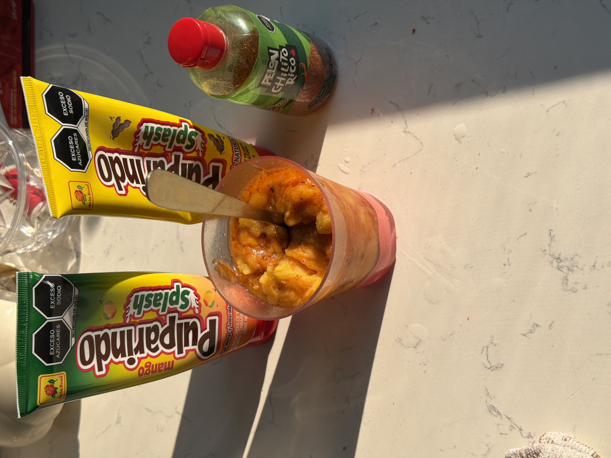 The image presents a top-down view of a snack arrangement on a light-colored countertop with subtle gray marbling, likely indoors. Predominantly, the composition includes two tubes of "Pulparindo Splash," one mango-flavored in green and the other tamarind-flavored in yellow. A bottle of "Pelon Pelo Rico" chili candy is also visible. The focal point is a plastic cup filled with what appears to be a slushy fruit mixture, partially consumed, with a silver spoon inserted. Shadows from an external light source suggest the photograph was taken during daylight. The location, city of Cuautla, Mexico, is not visually evident in the image.