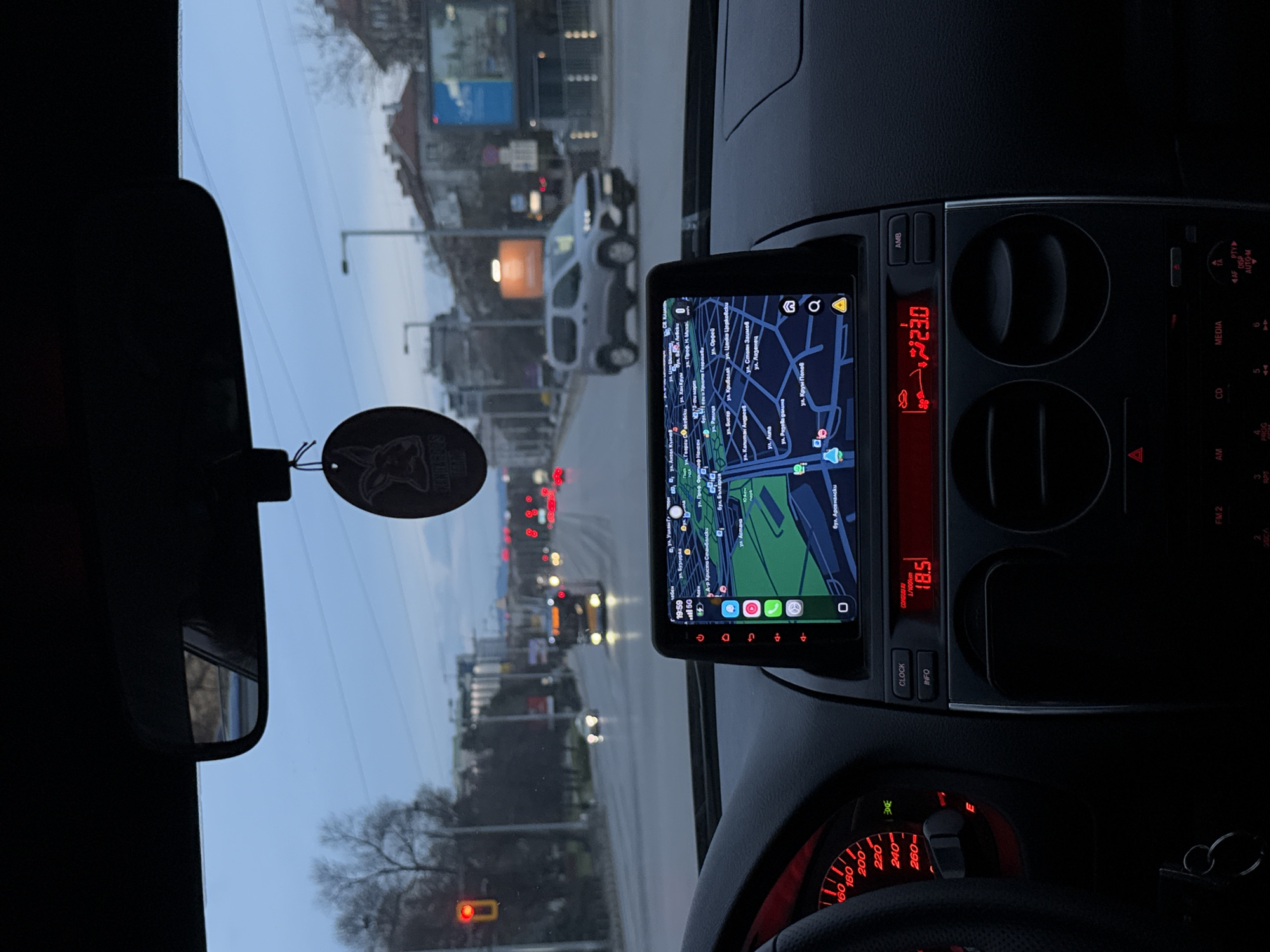 The image is taken from inside a vehicle, likely the driver's perspective, in the city of Sofia, Bulgaria. The dashboard is visible, with a GPS screen displaying a map of the area. The GPS screen shows streets such as "ул. Ангел Кънчев" and "бул. България," indicating the approximate location.
The car's dashboard also displays the time (19:59) and an outside temperature of 23 degrees Celsius. Other car features visible include the rearview mirror with an air freshener, the steering wheel, and the speedometer.
Outside the car, the street is visible with other vehicles, traffic lights, and buildings, suggesting an urban environment at dusk. The street is illuminated by headlights and streetlights.
