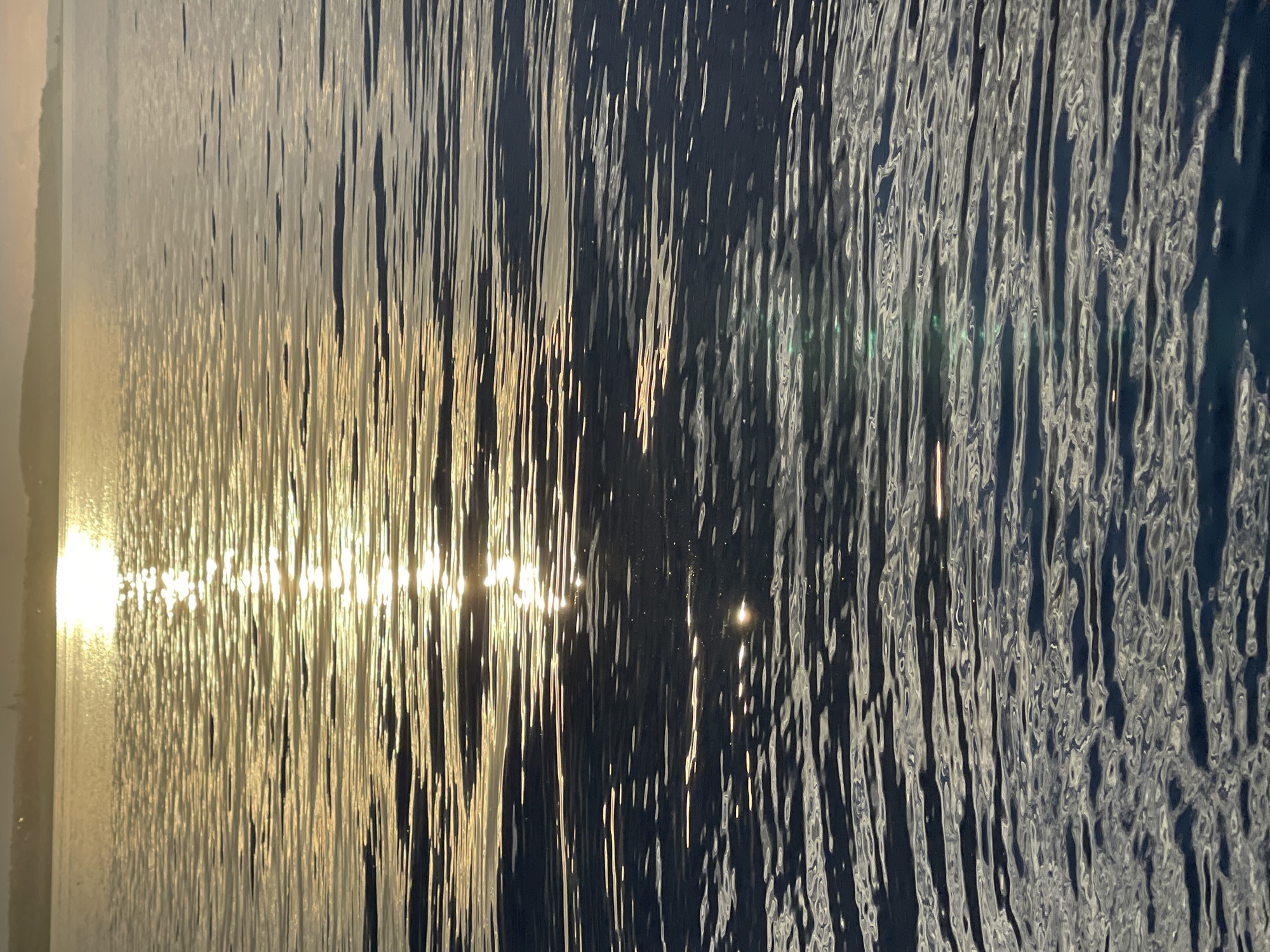 This image captures a close-up, abstract view of rippling water, likely a lake or ocean, at sunset. The setting sun, located on the far left of the frame, casts a bright, golden light that reflects intensely on the water's surface. These reflections create vertical streaks of shimmering light, interspersed with darker, rippled shadows. The water's surface is highly textured, with numerous crests and troughs creating a dynamic pattern of light and dark.
As the viewer's eye moves from left to right, the intensity of the sunlight diminishes, and the water appears darker, revealing a deep blue hue. The reflections on the right side are more subdued, appearing as small, scattered glints of light. The overall impression is one of tranquility and natural beauty, with the interplay of light and shadow on the water evoking a sense of movement and depth. The time of day is clearly indicated by the golden light of the setting sun, suggesting either early morning or late evening. There are no visible people, objects, or text in the image. The focus is entirely on the abstract patterns formed by light and water.