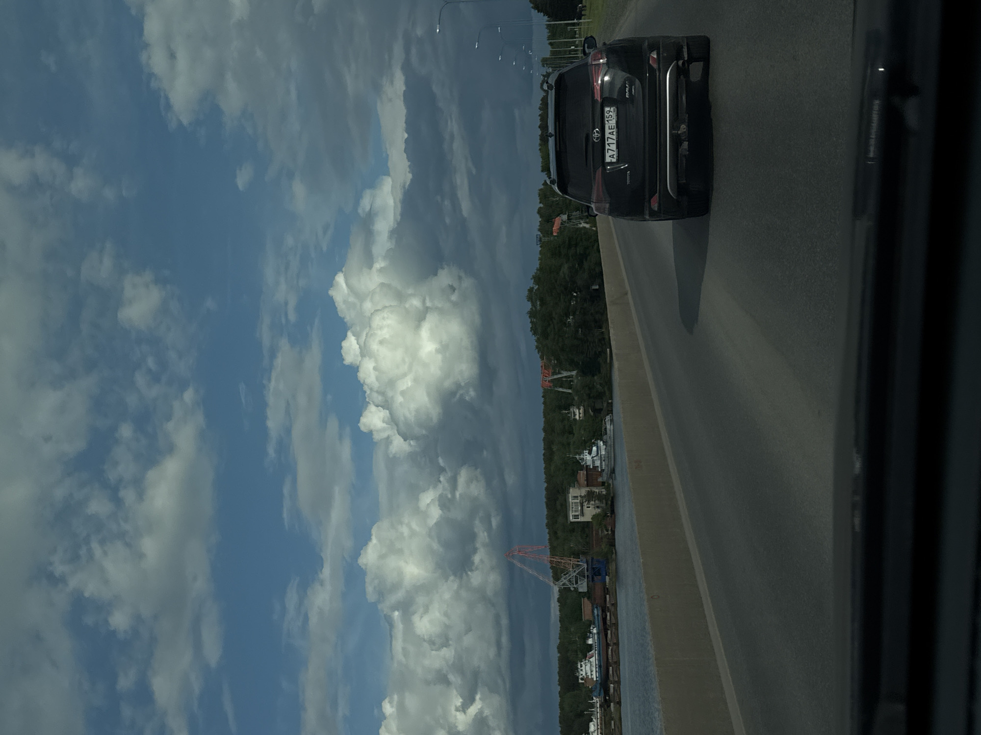 The image shows a view from a vehicle traveling on a bridge near Chaikovsky, Russia. To the right, a black Toyota RAV4 with license plate "A717AE159" is visible on the road. The background reveals a landscape with trees, buildings, and an industrial crane next to a body of water. The left side of the frame is dominated by a blue sky filled with white cumulus clouds. The bridge appears to be made of concrete.